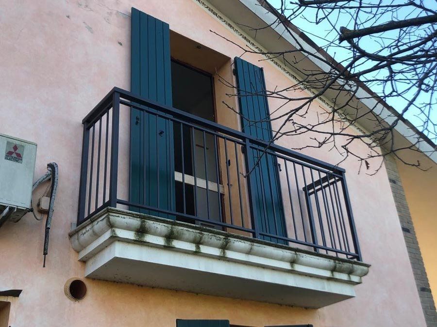 Balcone con persiane verde scuro, ringhiera nera ed esterno in stucco rosa.