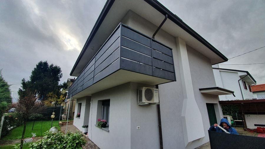 Casa a due piani con balcone caratterizzato da pannelli grigio scuro, sullo sfondo di un cielo nuvoloso.