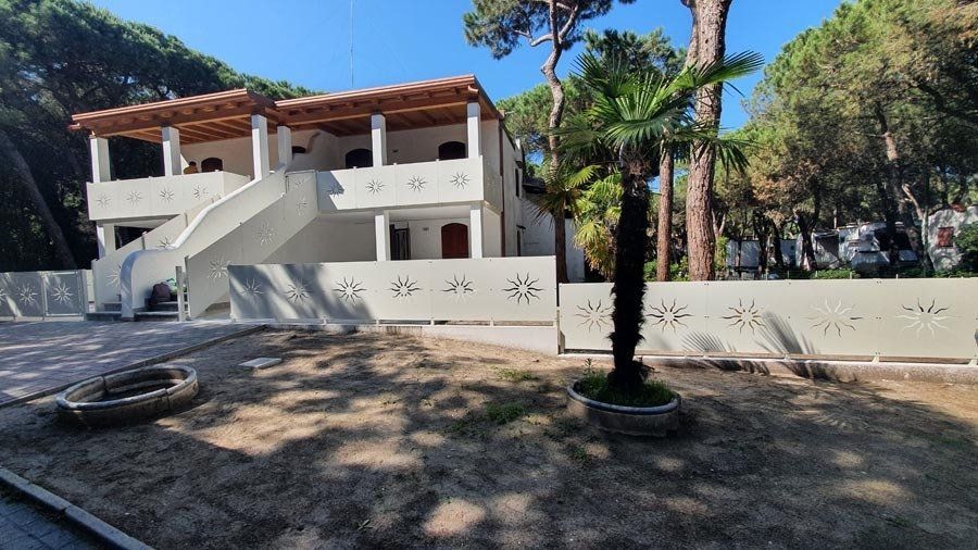 Edificio a due piani di colore chiaro con balconi, una scala bianca e una recinzione decorativa; palma di fronte.