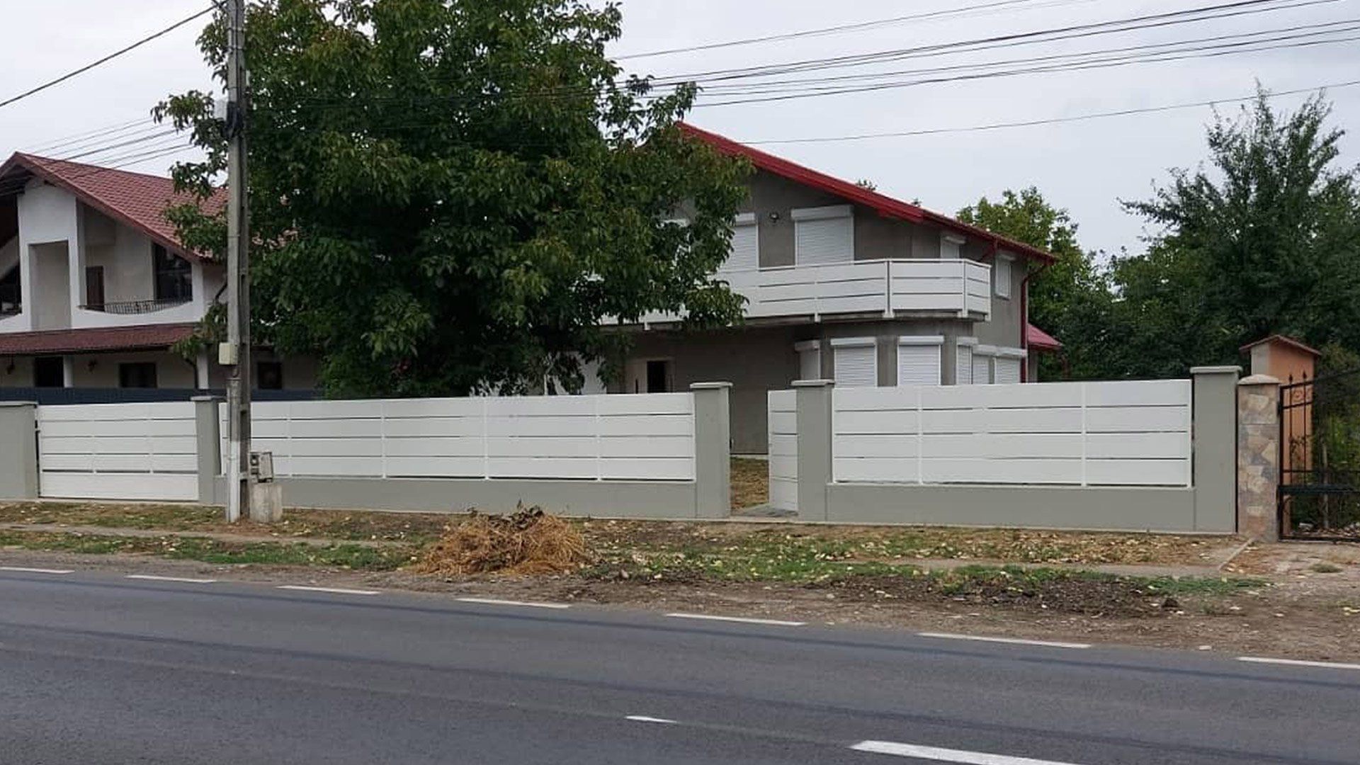 Una casa a due piani con una staccionata bianca lungo la strada. La casa ha il tetto rosso e il rivestimento grigio.