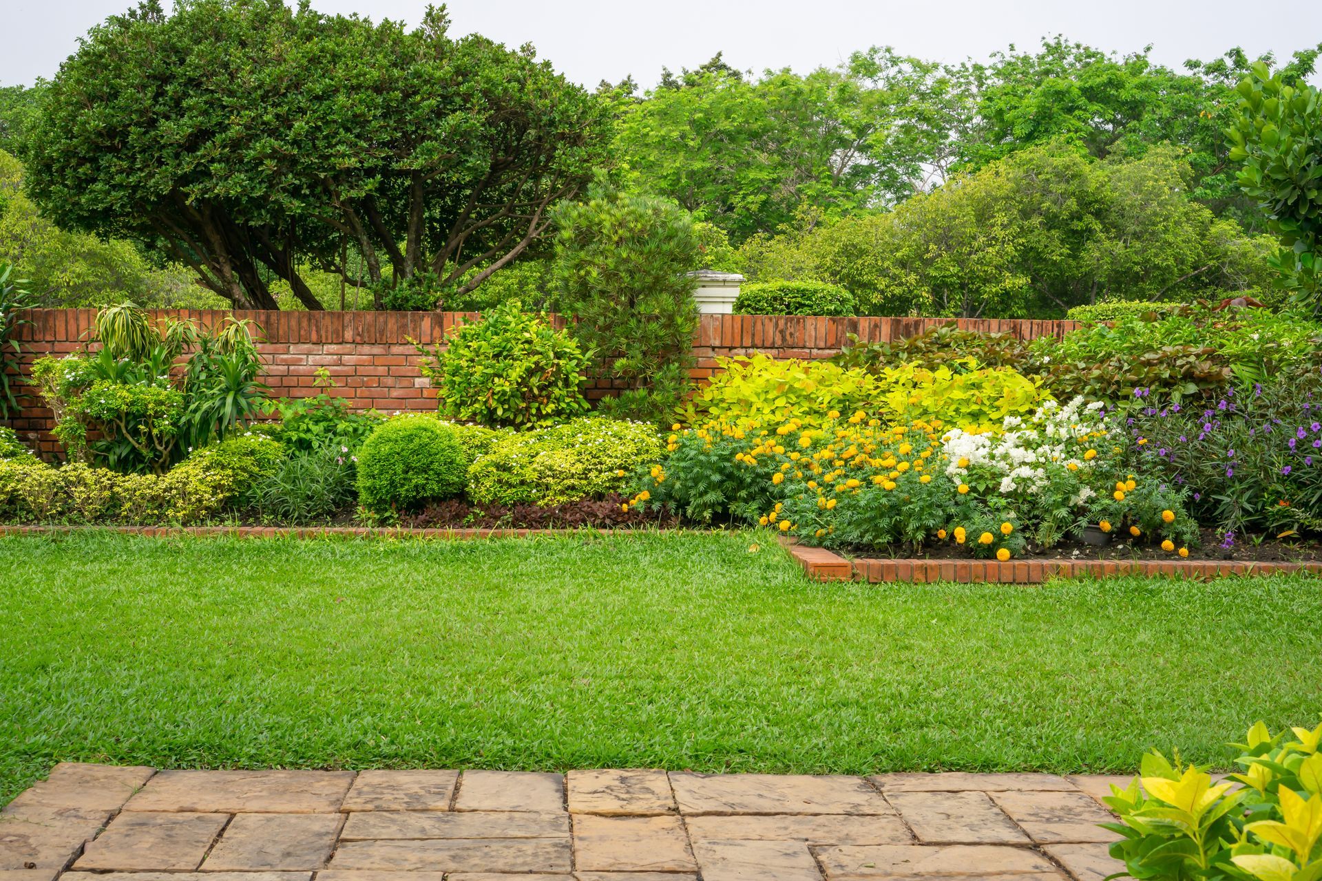 Green lawn with a colorful garden bed in front of a brick wall, lush trees in background.