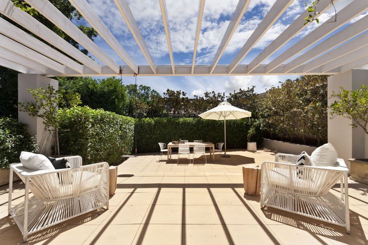 A patio with chairs , a table and an umbrella under a pergola.