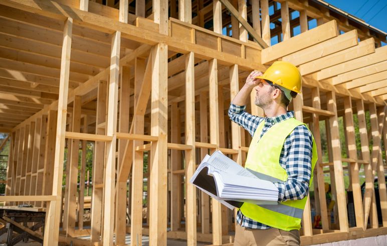 A construction worker is standing in front of a wooden house under construction.