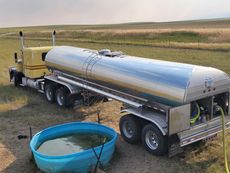 Yellow tanker truck filling a blue water trough in a grassy field.