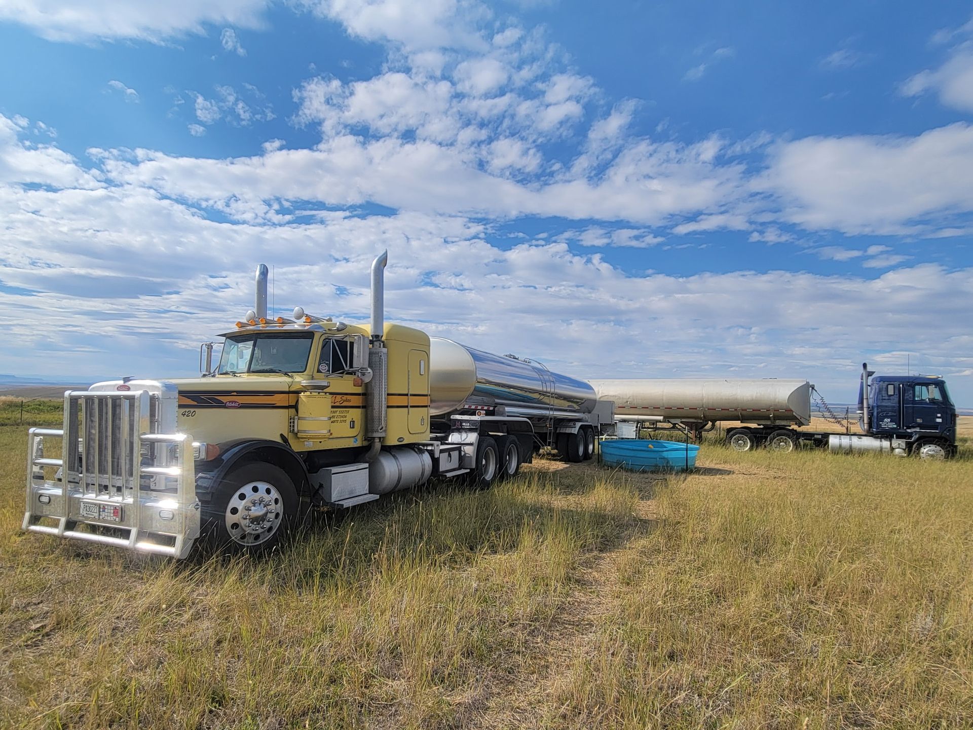 Yellow semi-truck with a tank trailer in a field under a blue sky with clouds. Another blue truck is in the background.