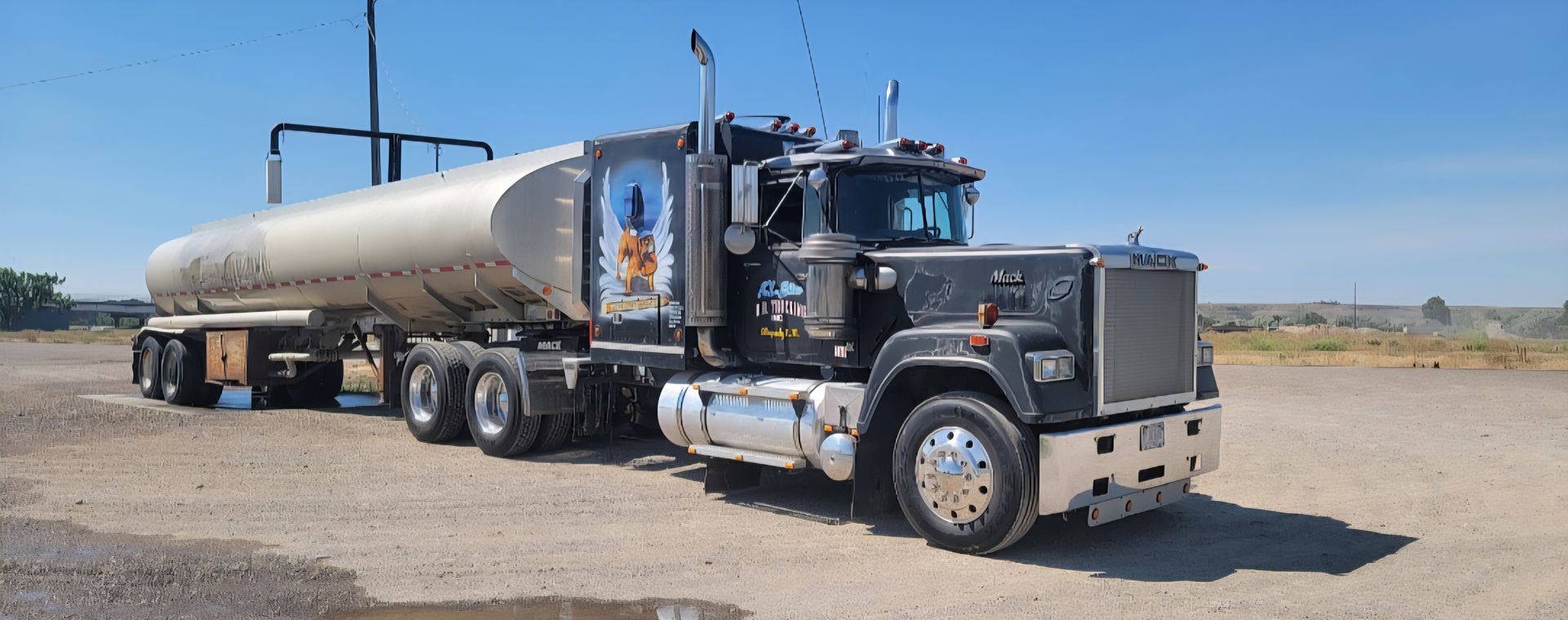 A black tanker truck parked on a gravel surface under a clear blue sky.