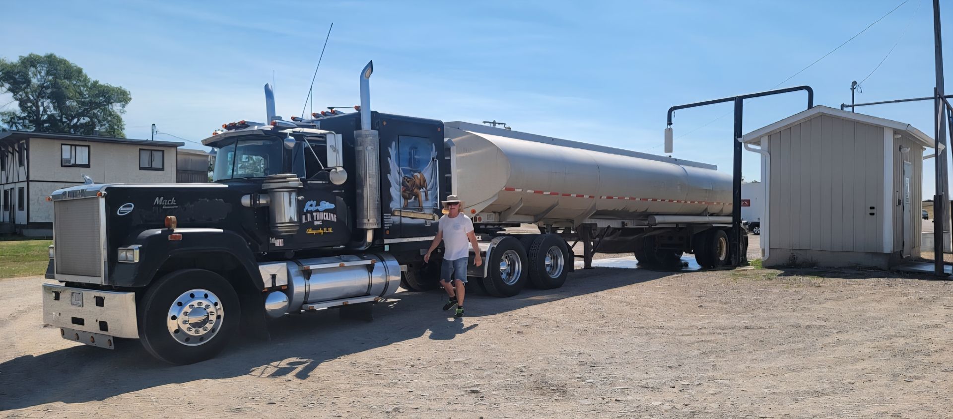 Black tanker truck parked next to small building, man stands near truck, bright blue sky.
