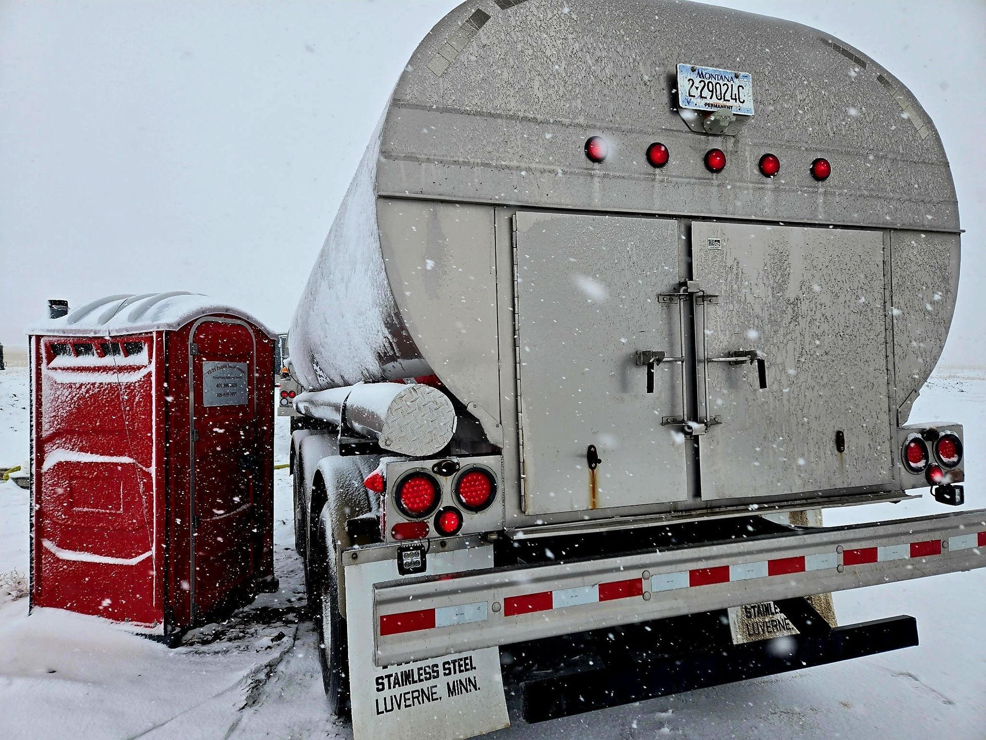 Red portable toilet beside a large silver tanker truck in snowy conditions.