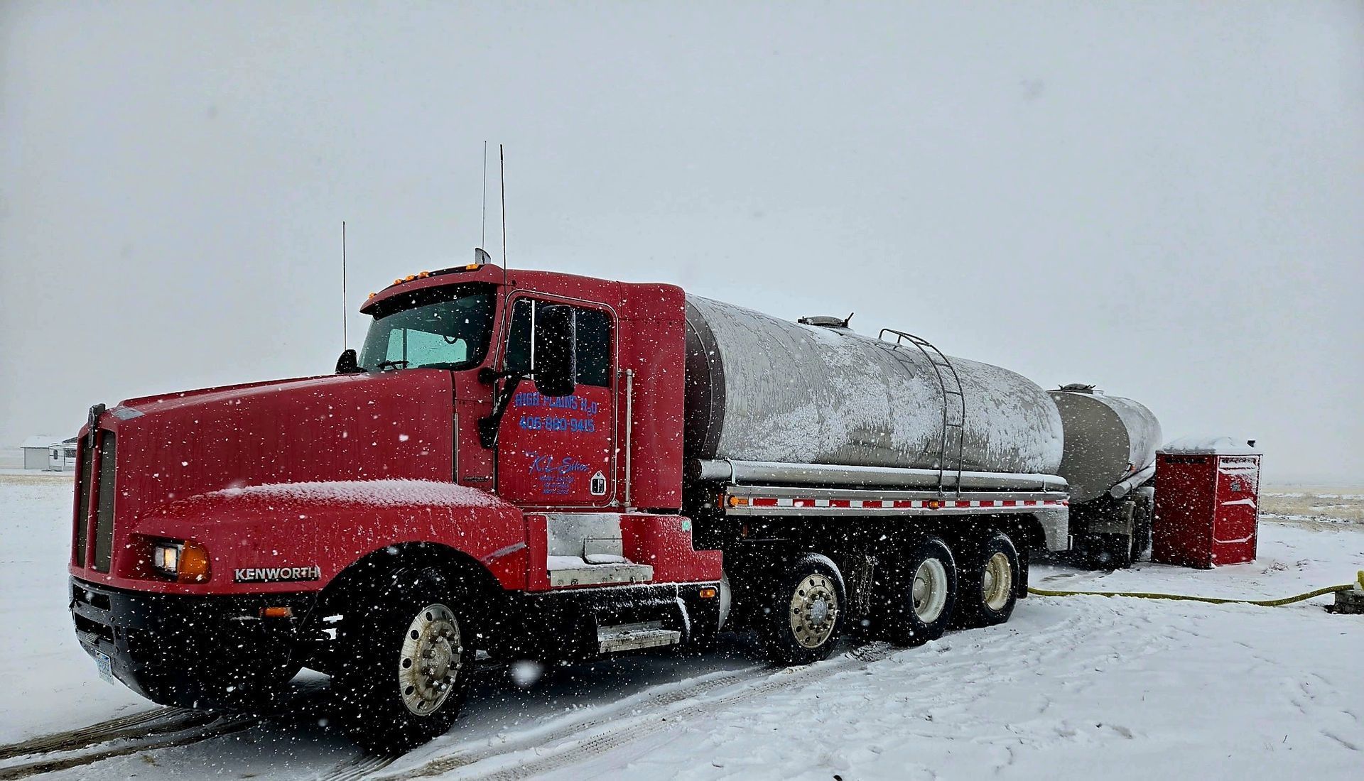 Red tanker truck in snowy conditions, parked next to a tank, in an open field.
