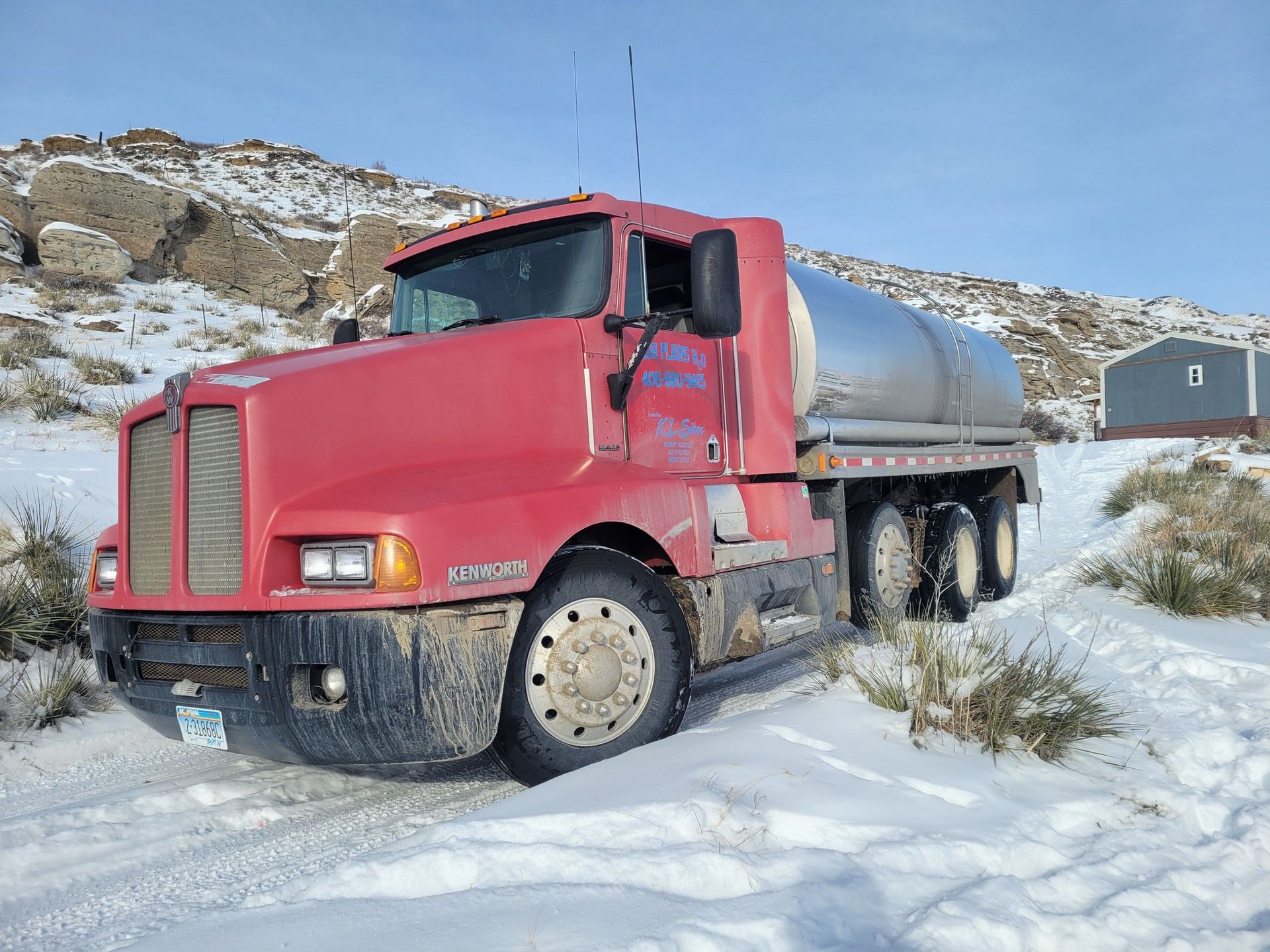 Red water tanker truck driving on a snowy, dirt road near a small building and rocky hills.