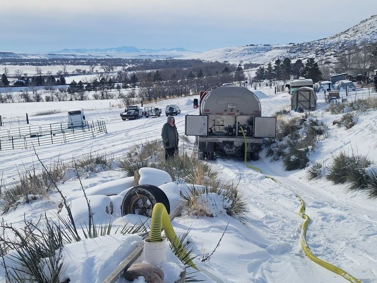 Snowy landscape with a truck and person near a yellow hose, with hills and vehicles in the background.