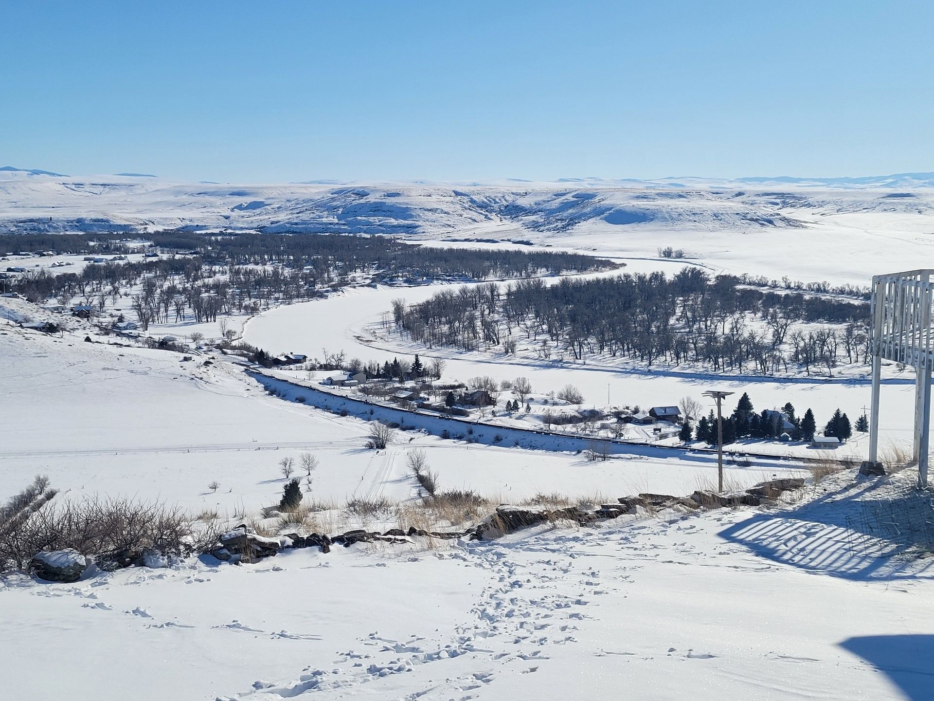 Snowy landscape with trees, a river, and buildings under a clear blue sky.