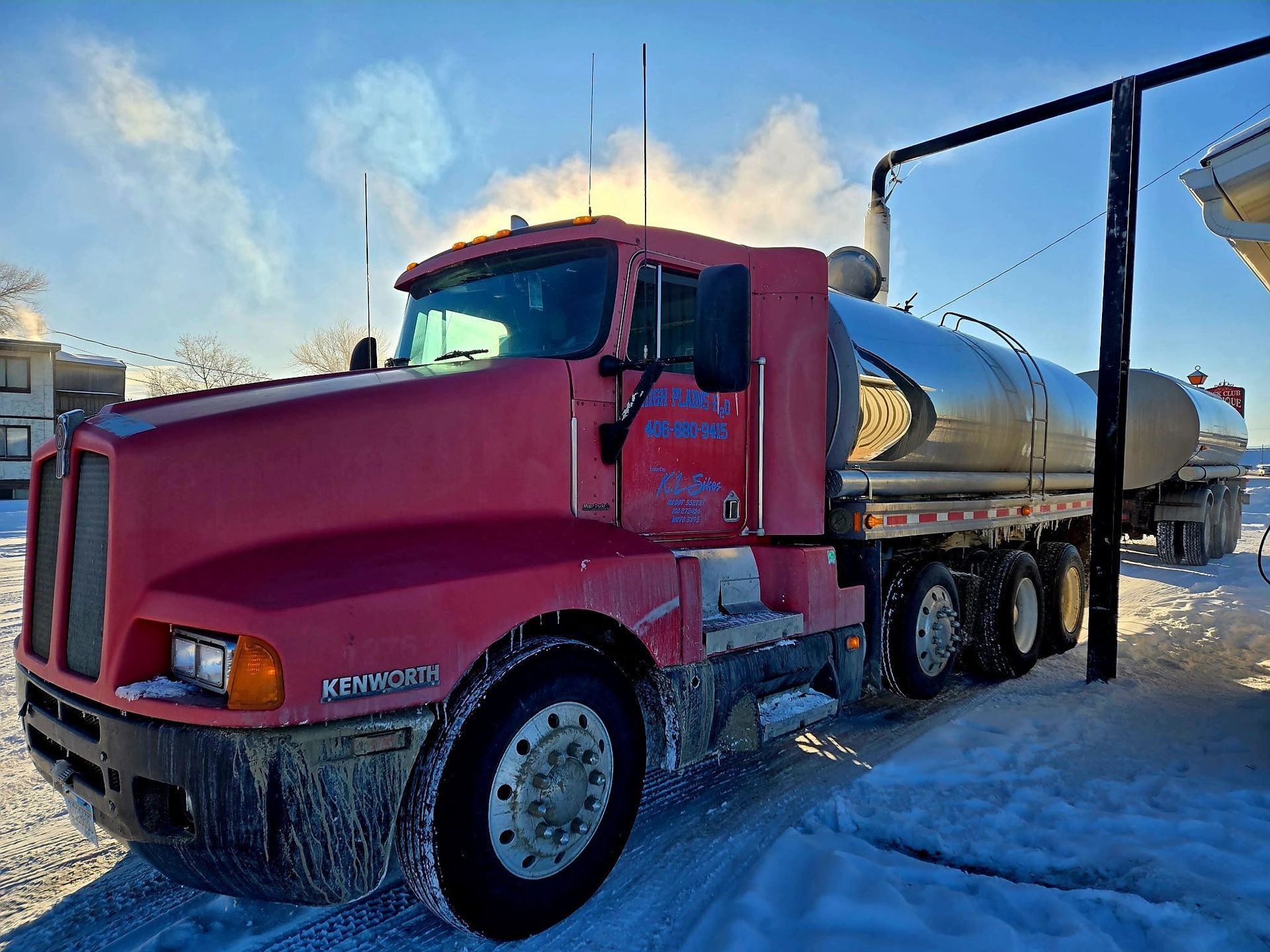 Red Kenworth tanker truck parked in snow, sun shining.