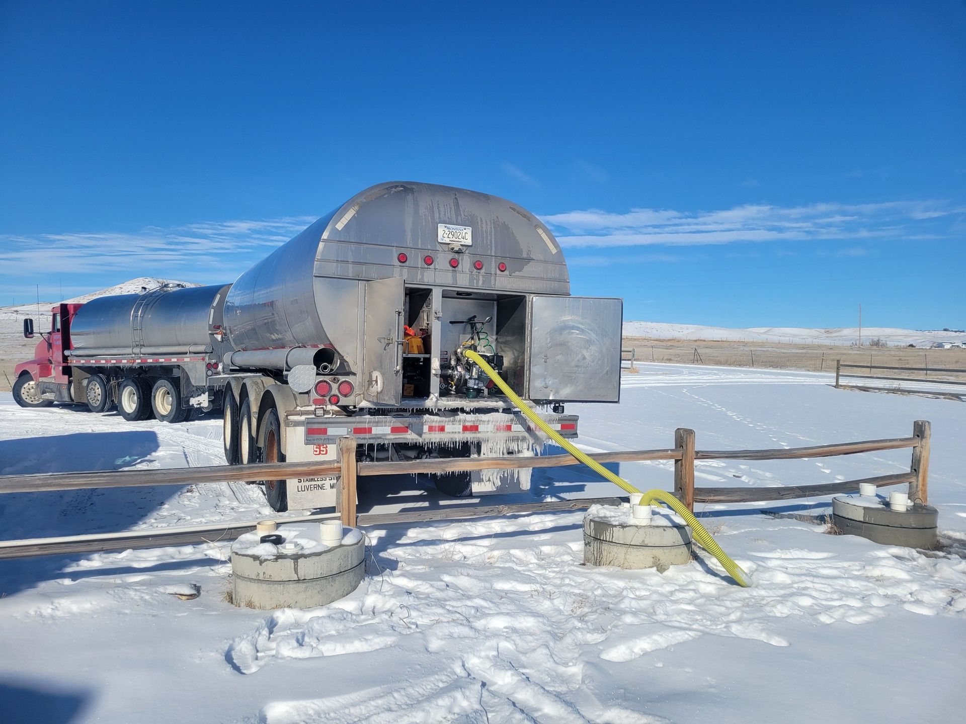 Fuel tanker truck filling a tank in a snowy, rural setting. Yellow hose extended between tanks.