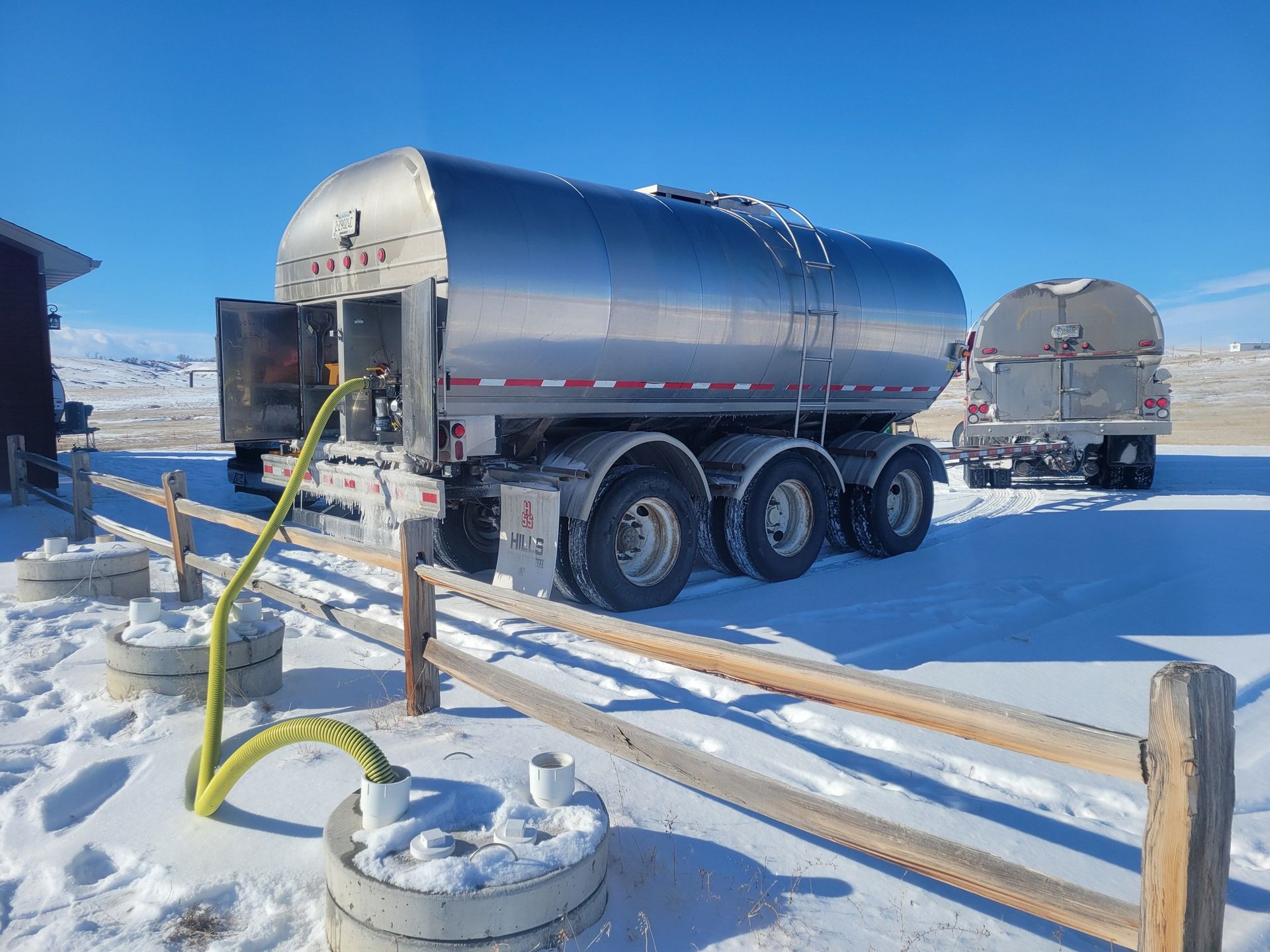 Milk tanker truck parked in snowy field, connected to a milk storage tank by a hose.