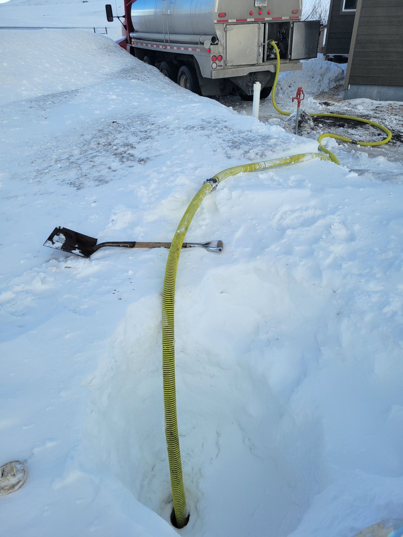 A yellow hose extends from a water truck across a snowy trench. A shovel rests nearby.