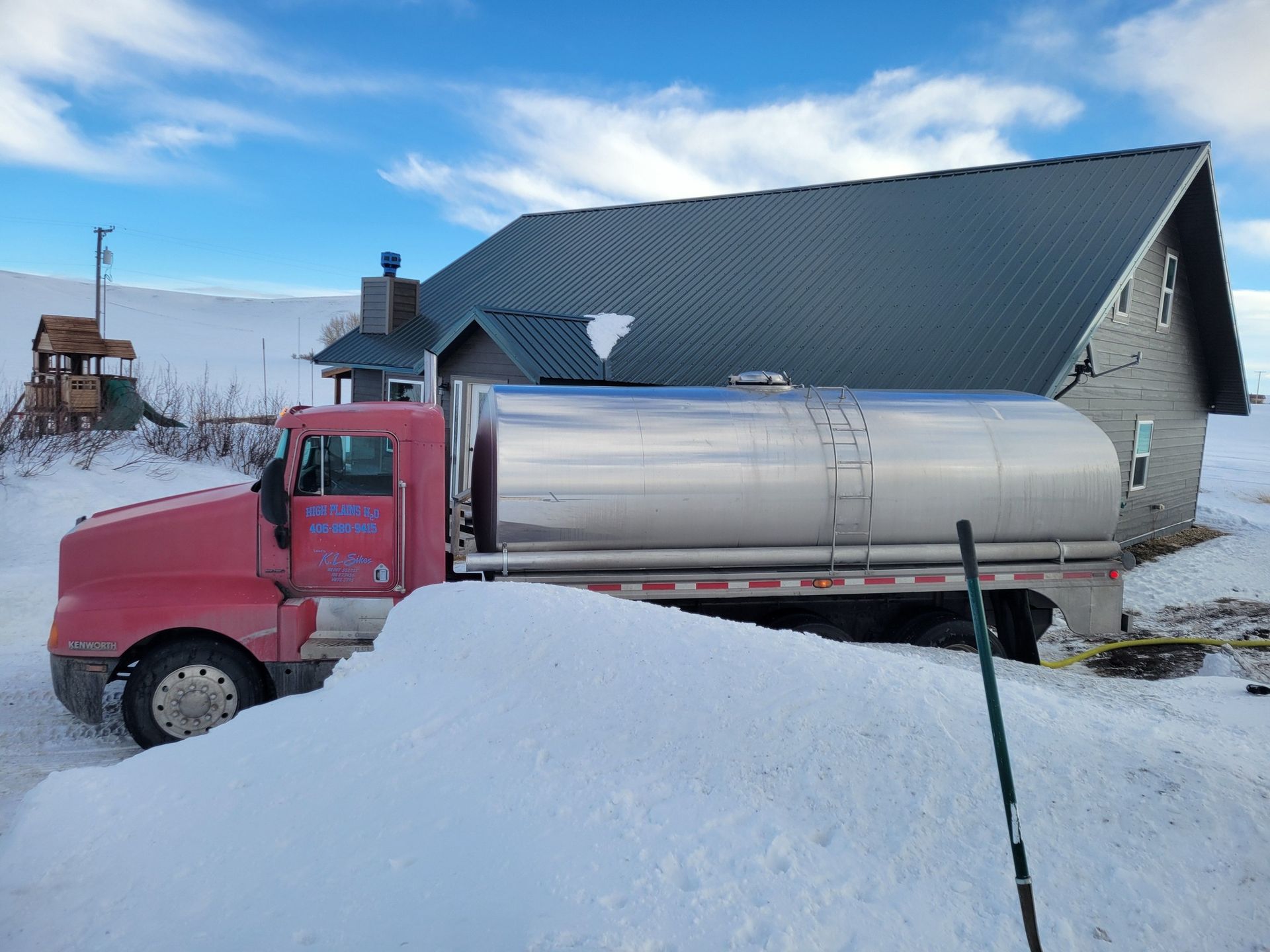 Red milk truck parked near a gray building in a snowy outdoor setting under a blue sky.