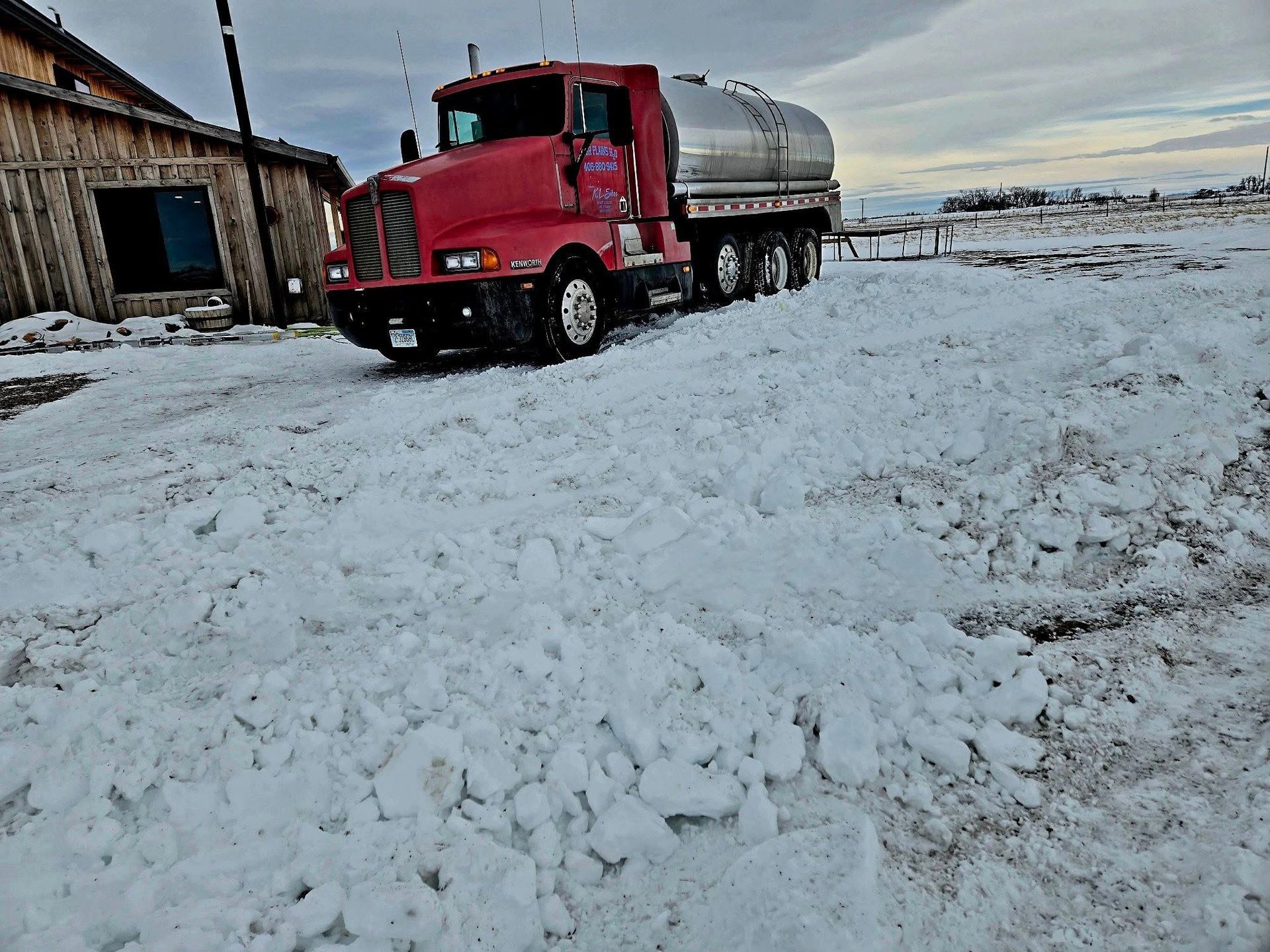 Red tanker truck driving through snow near a wooden building in a rural setting.