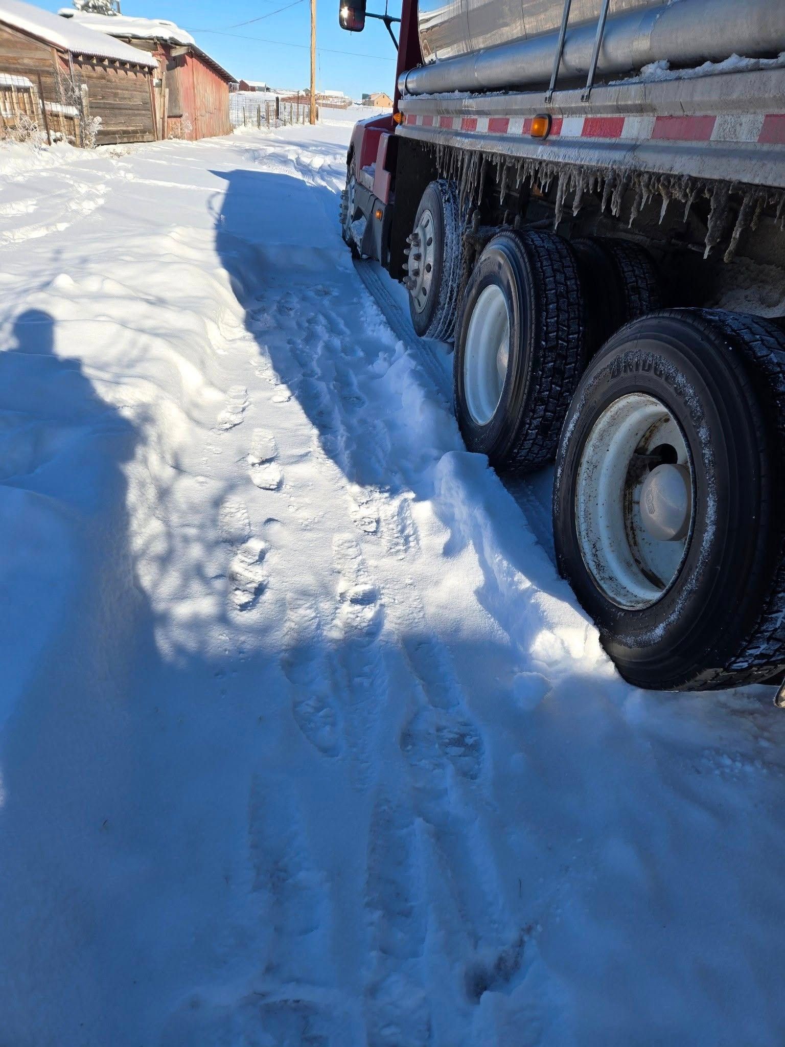 Semi-truck on a snow-covered road with tracks. Buildings visible in the background on a sunny day.