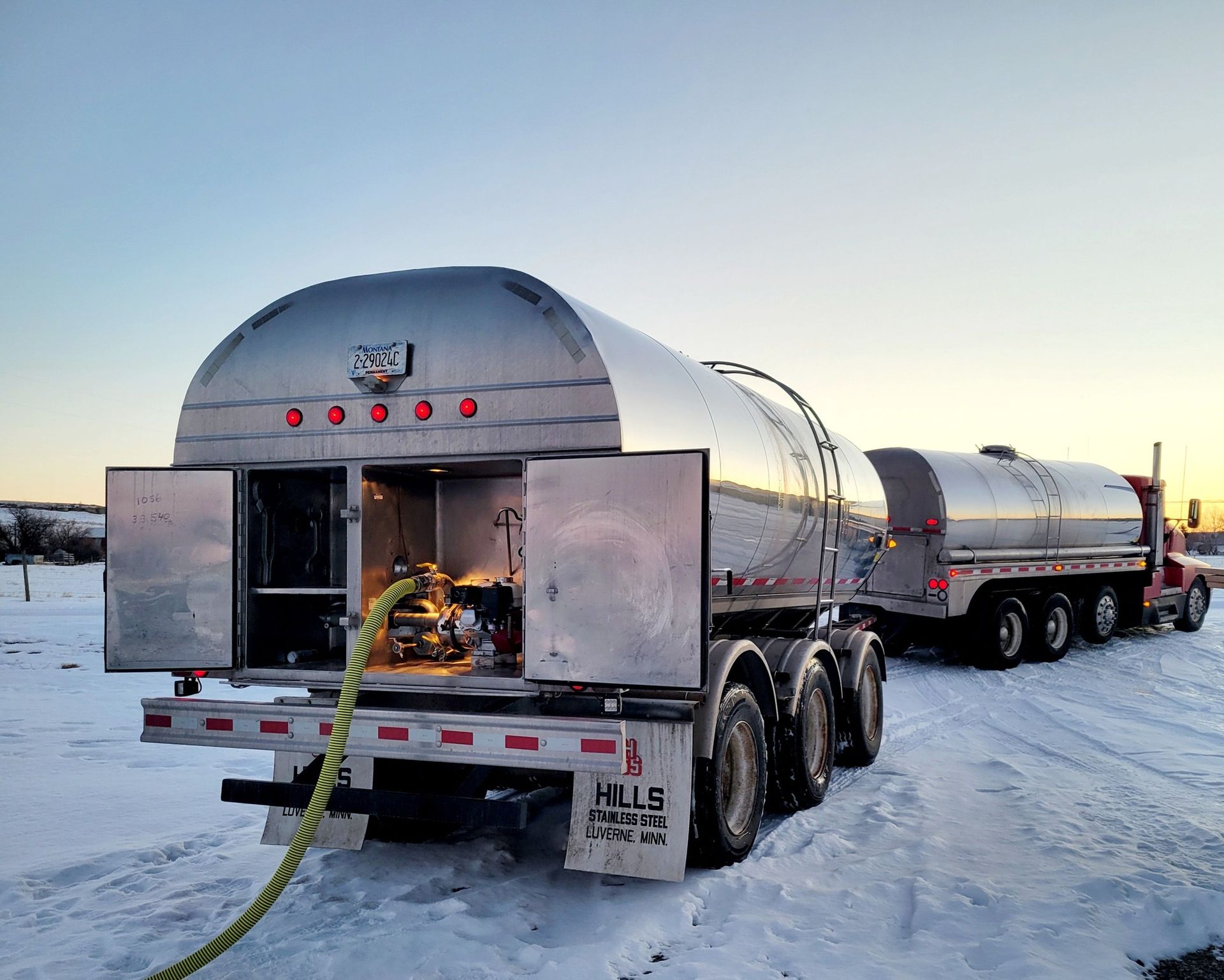 A shiny silver tanker truck with open back doors, yellow hose, and another truck in a snowy field at sunset.