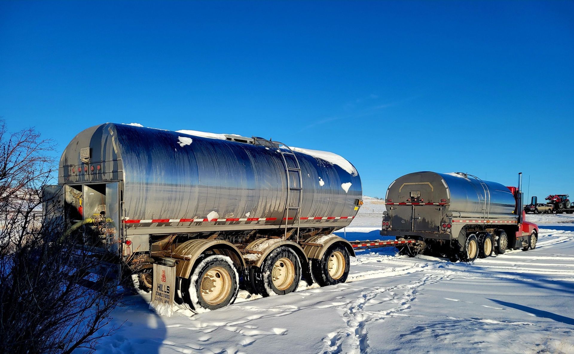 Tanker trucks in snowy field, one pulling the other; bright blue sky.