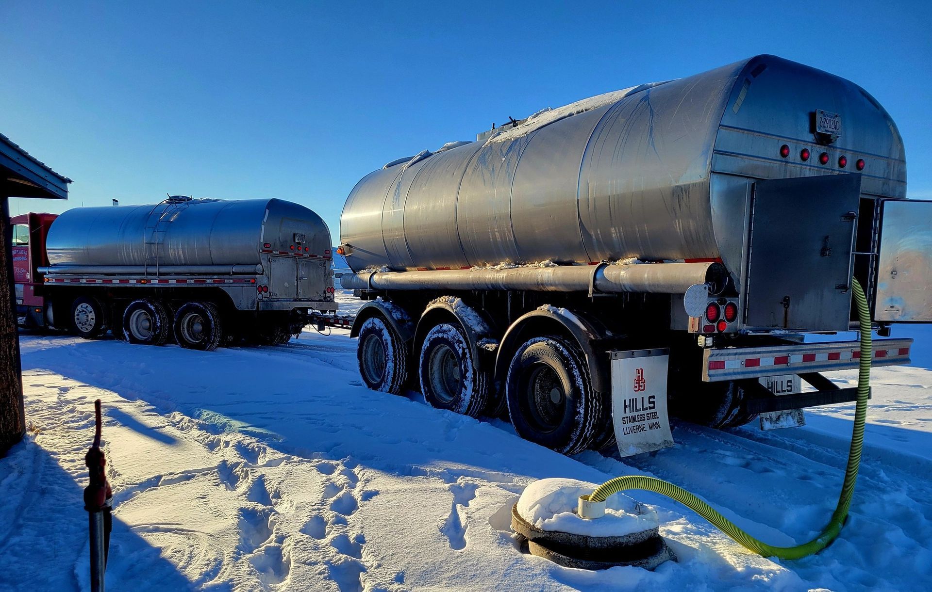 Two tanker trucks parked in snow; a green hose connects one truck to a covered pipe in the ground.