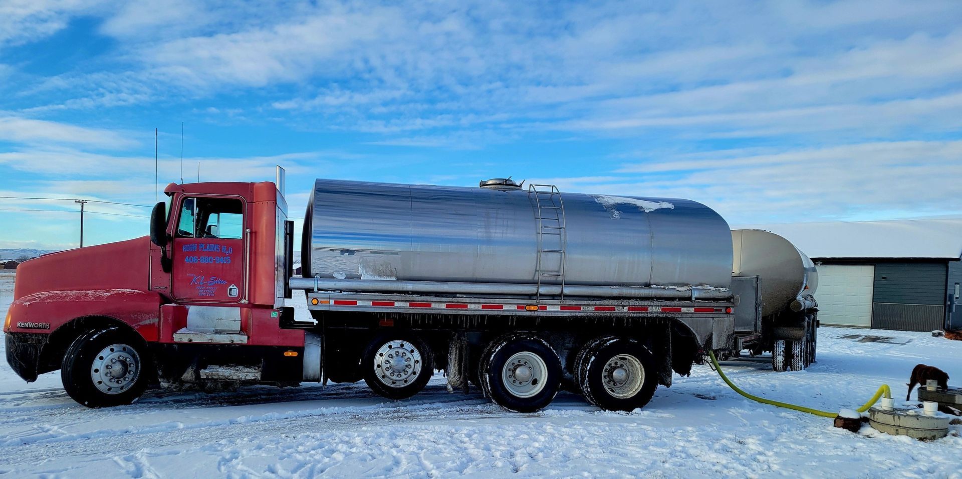 Red tanker truck parked in snowy landscape, hose connected to another tank, blue sky.