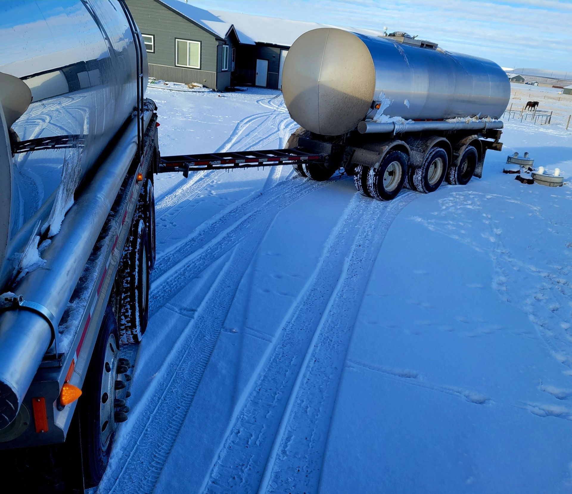 Truck pulling a silver tank trailer in a snowy yard; a house is in the background.
