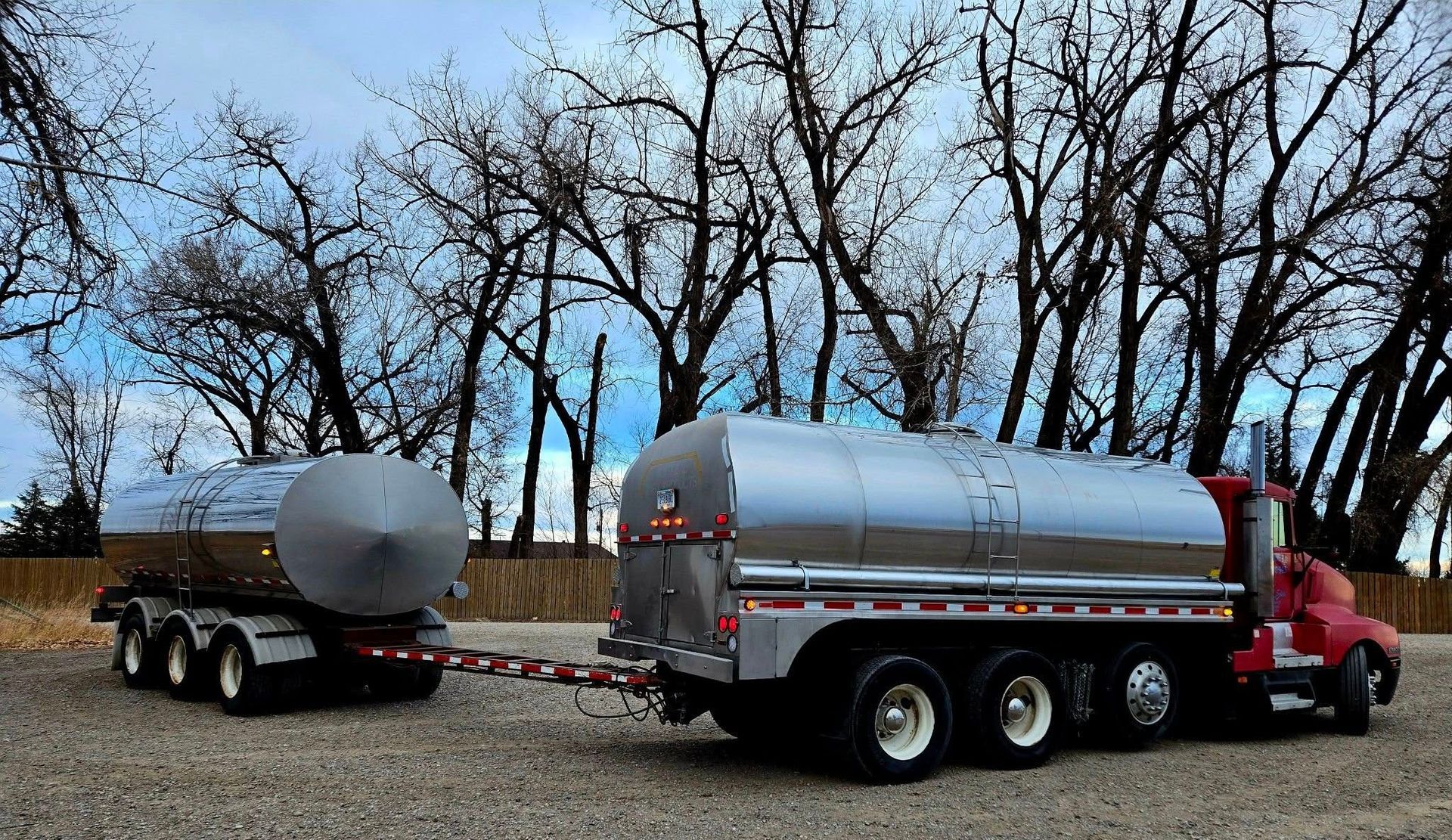 Red semi-truck pulling a silver tanker trailer and a smaller tanker, parked on gravel, trees in background.