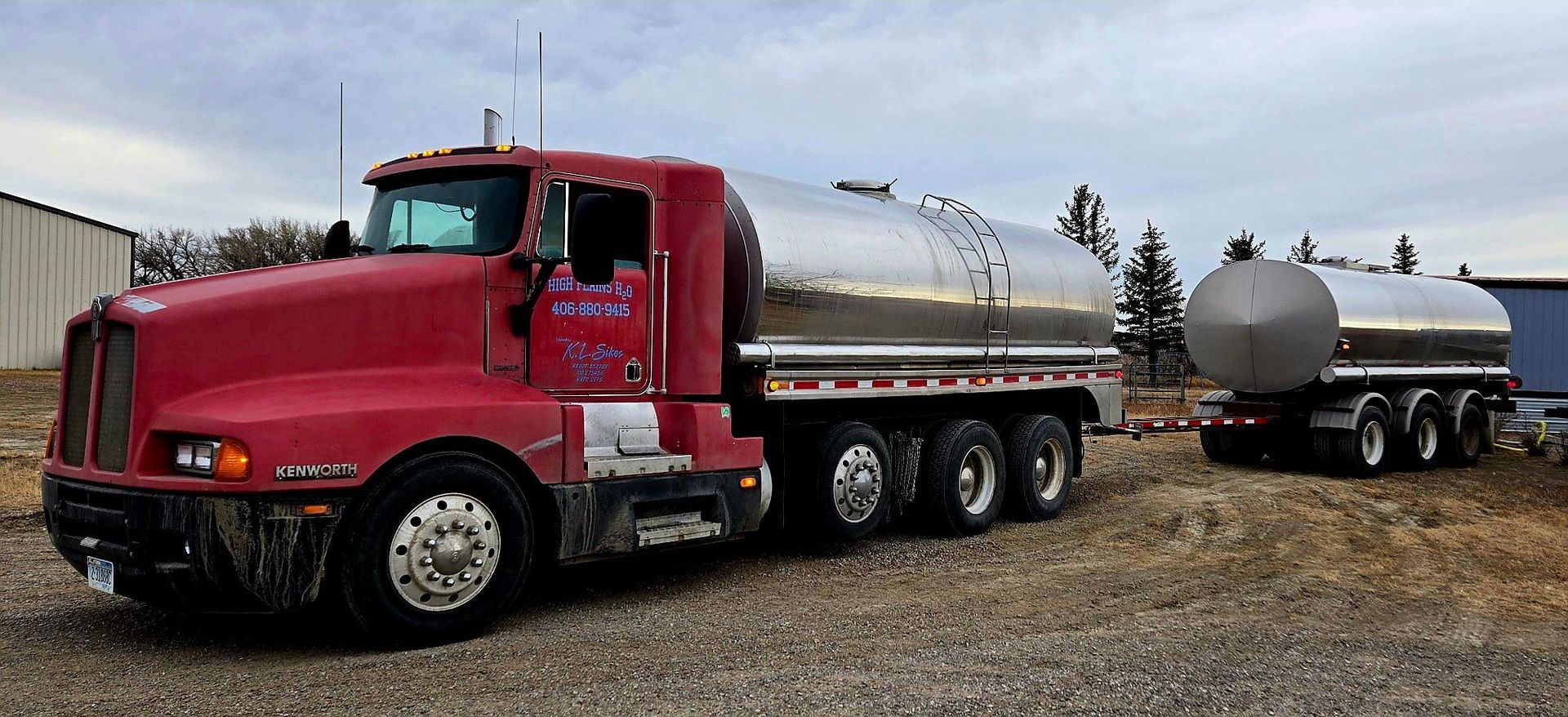 Red truck with tanker trailer parked on gravel, under cloudy sky.