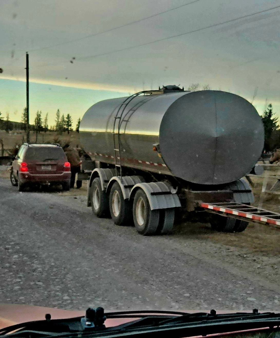 A large silver tanker trailer blocks a gravel road; a red car waits behind it.