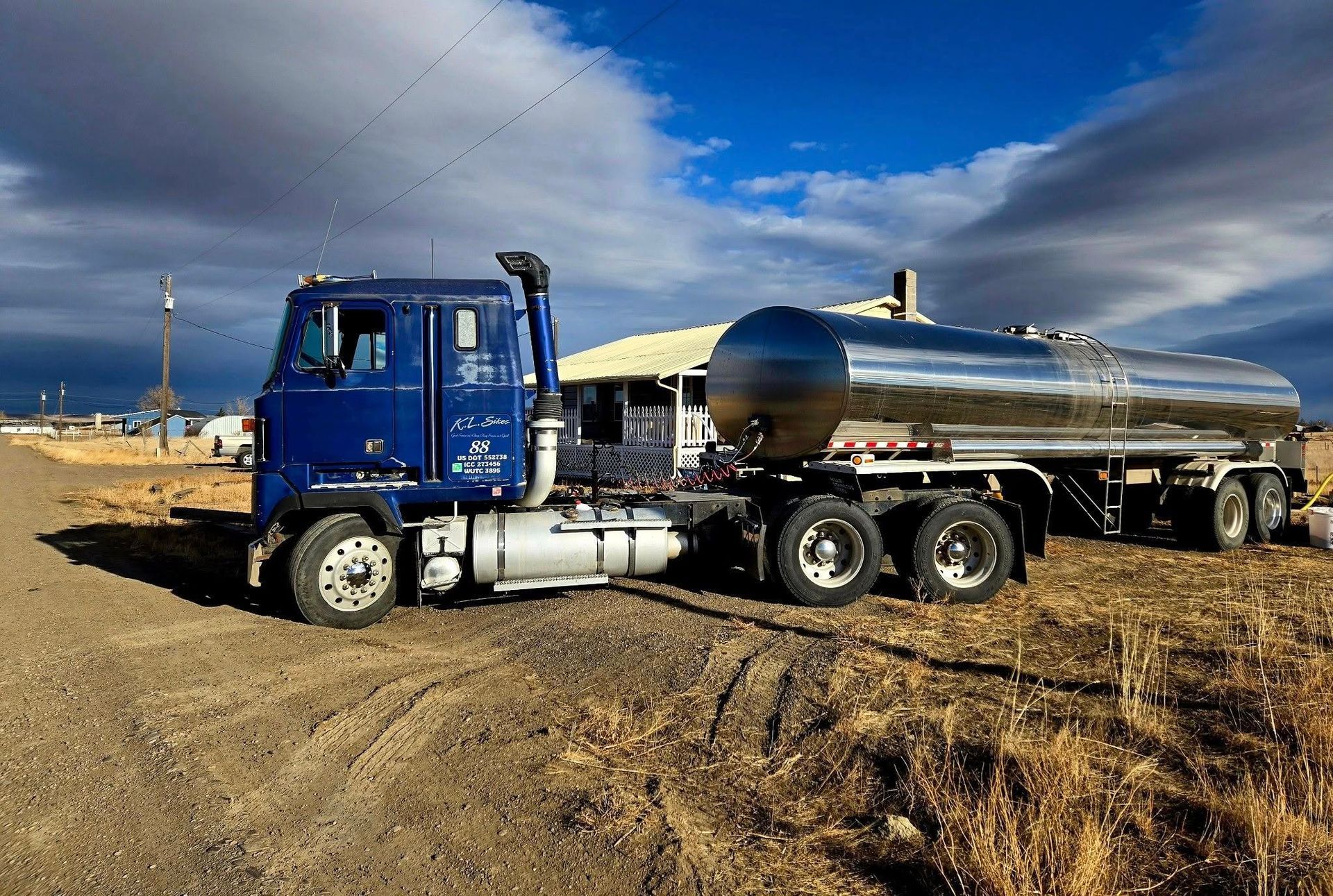 Blue tanker truck parked on a dirt road with a silver tank. Cloudy sky and small building in the background.