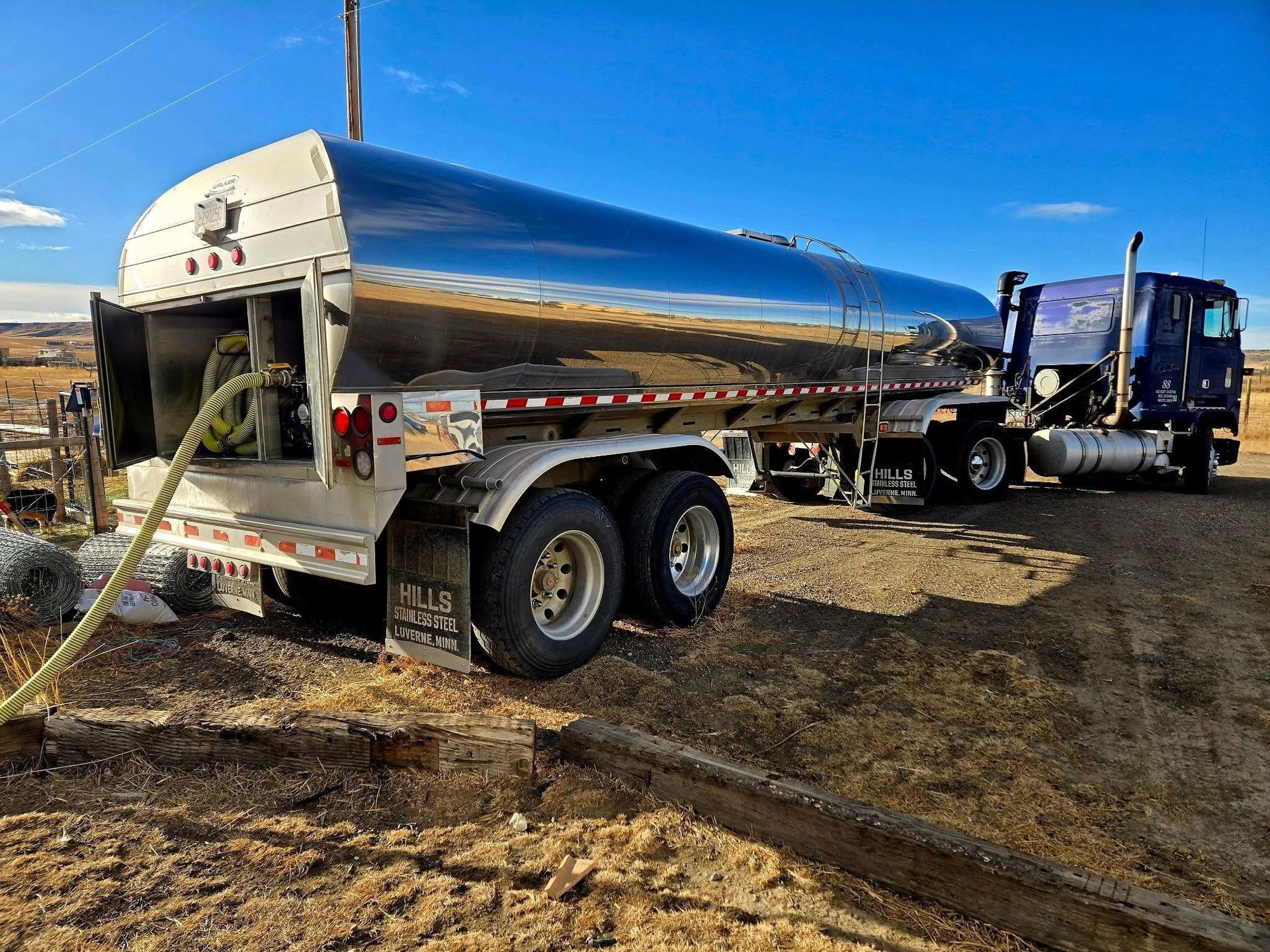 Fuel tanker truck in a field, connected to a hose; shiny silver tank reflects the sky.