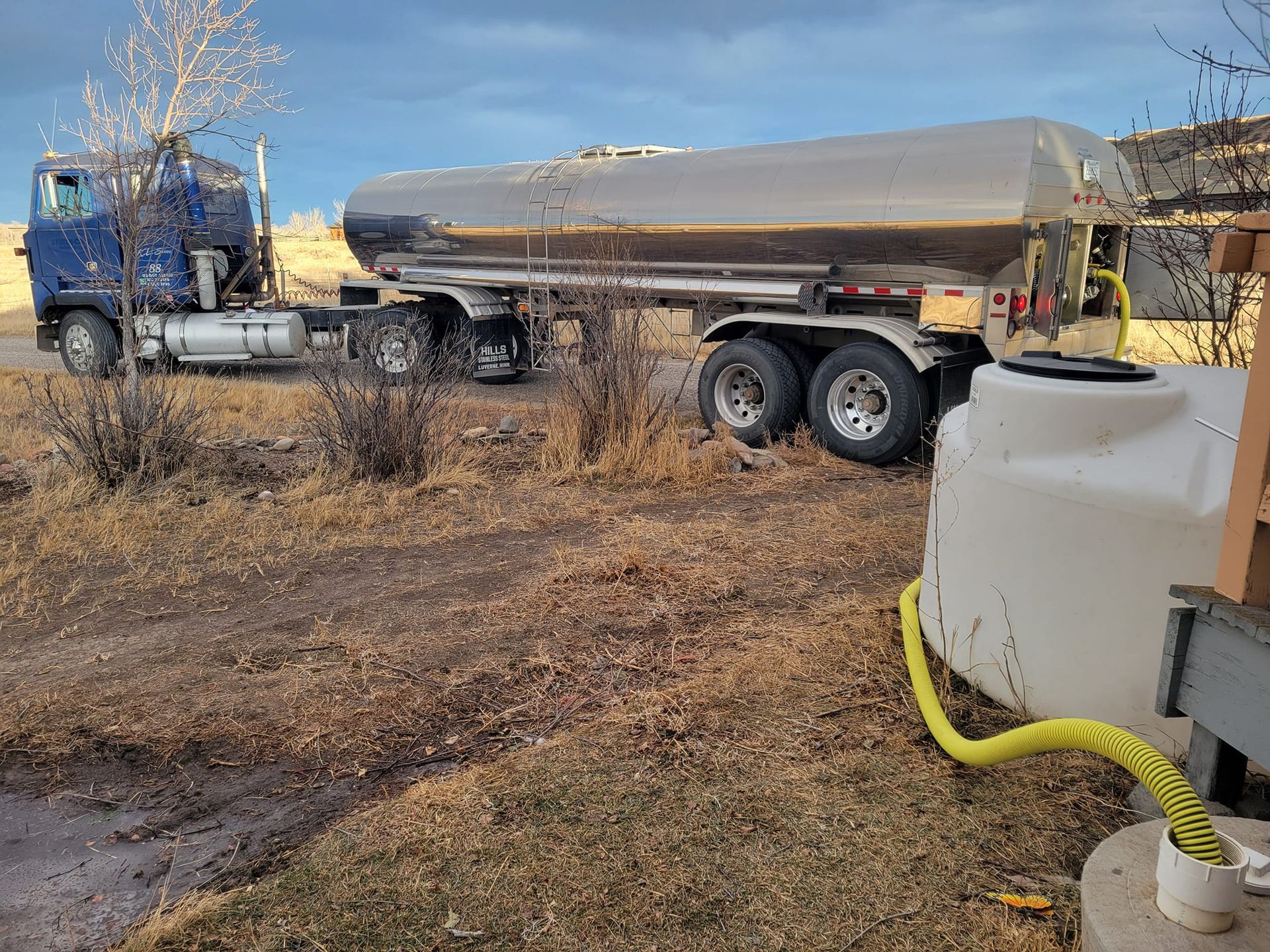 Blue tanker truck filling a white water tank with a yellow hose on brown grass under a cloudy sky.