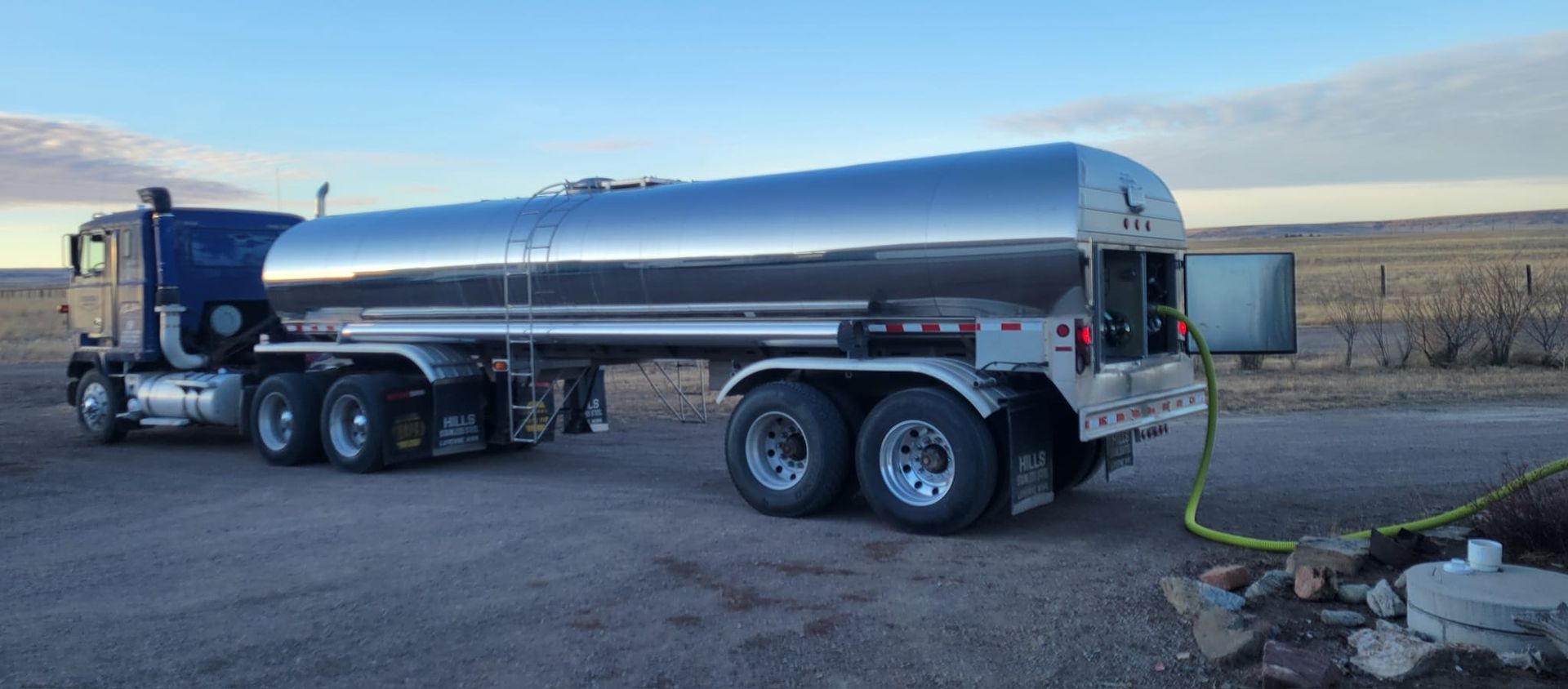 A tanker truck is parked on a gravel road, with a hose attached to a tank in a rural setting.