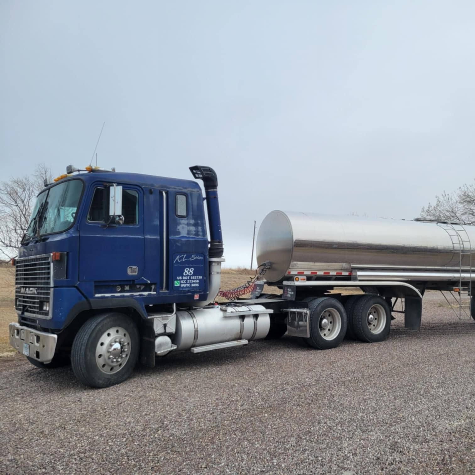Blue semi-truck with a silver tanker trailer parked on gravel under a cloudy sky.