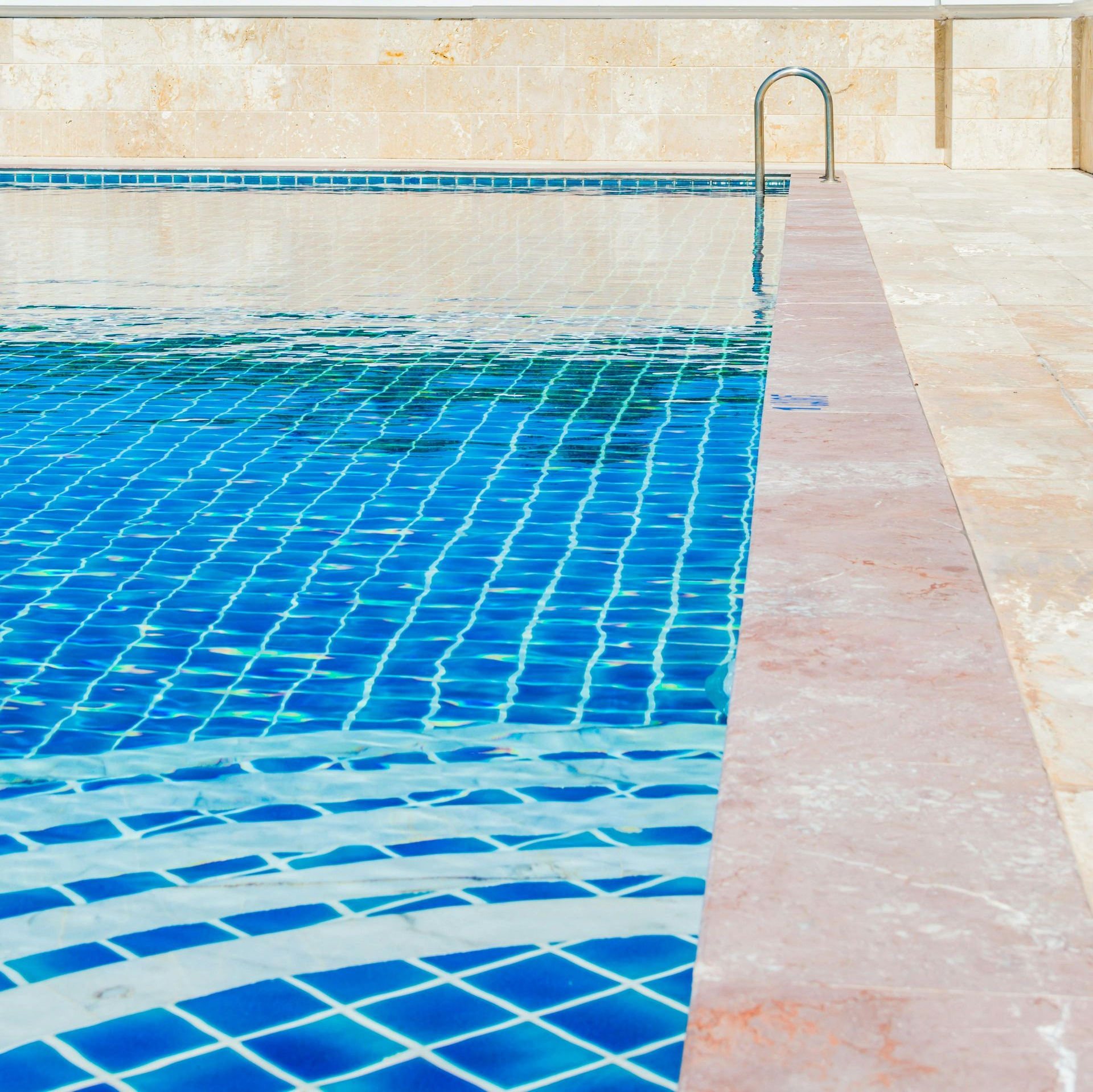 Swimming pool with blue tiled bottom, edge and handrail. Beige deck and wall.