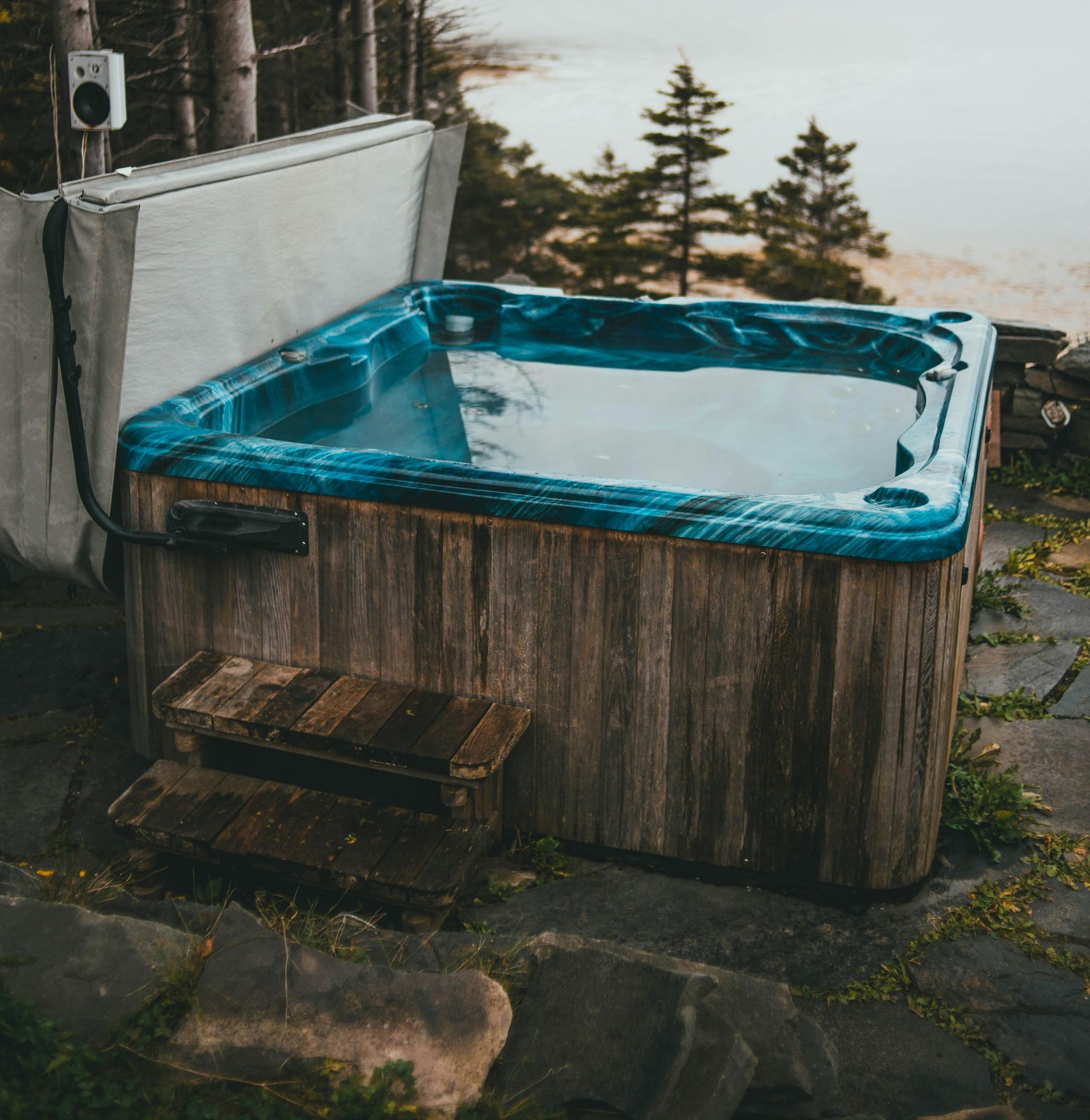 Hot tub with wooden exterior, next to rocky ground, water, and trees.