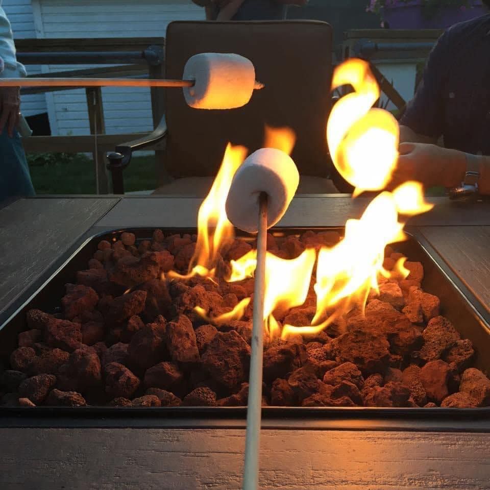 Marshmallows roasting over a fire pit. Close-up, two people hold marshmallows on sticks, flames, and lava rocks visible.