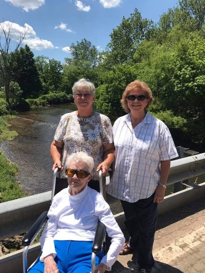 Three women on a bridge over a stream; one in wheelchair, two standing, bright sunny day.