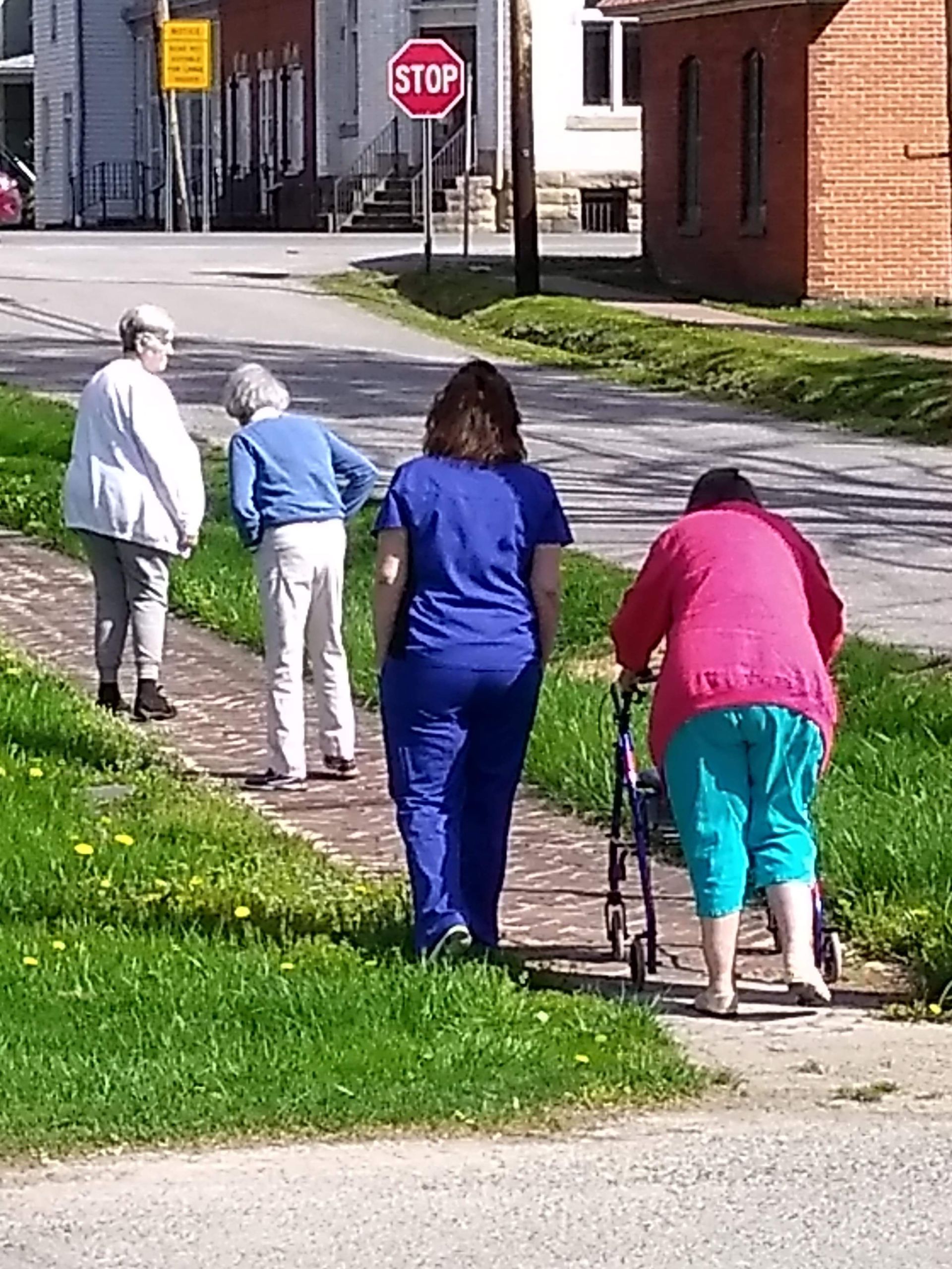 Four people walking on a sidewalk next to grass and a street. One uses a walker. Stop sign visible.