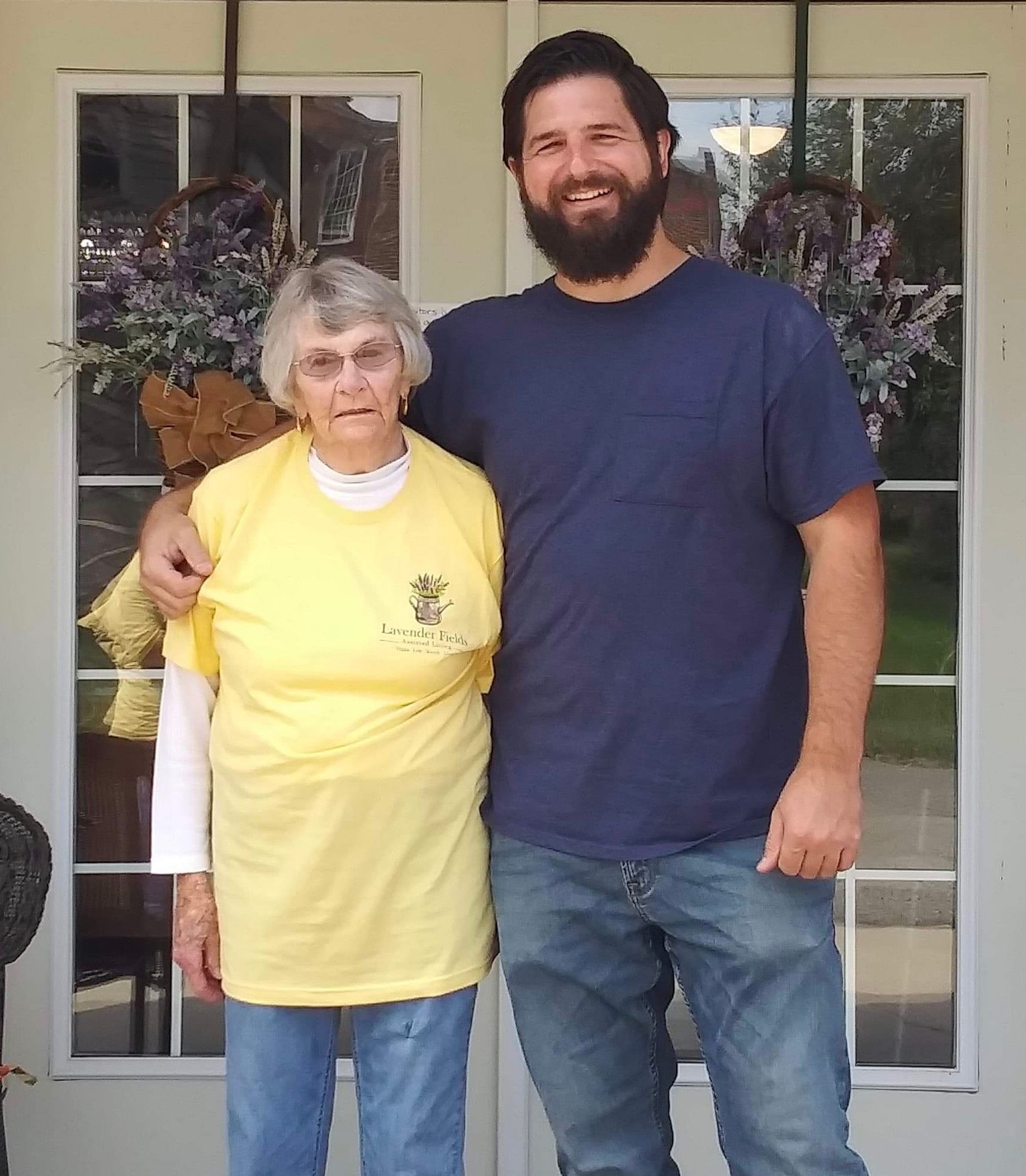 A man with a beard and an elderly woman stand together on a porch. The man has his arm around her.