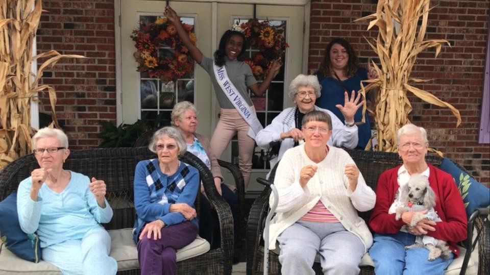 Group photo: Seniors with a woman holding a sash, posing outside a brick building with fall decor.