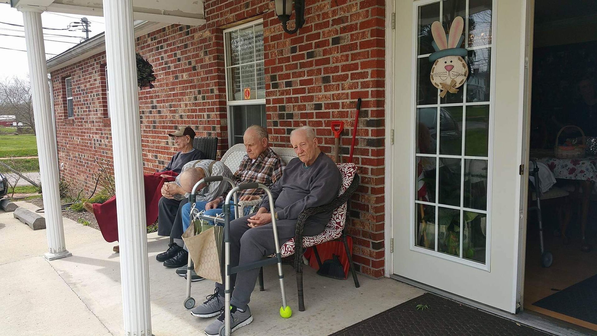 People on a porch; one using a walker. Brick building, white columns, and a door with a bunny wreath.