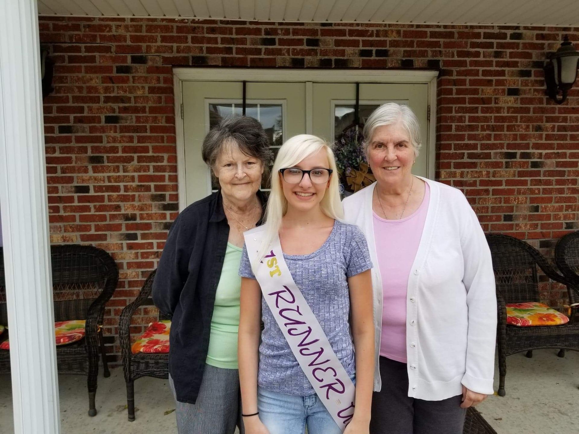 Three women standing on a porch in front of a brick building; young woman wearing a 