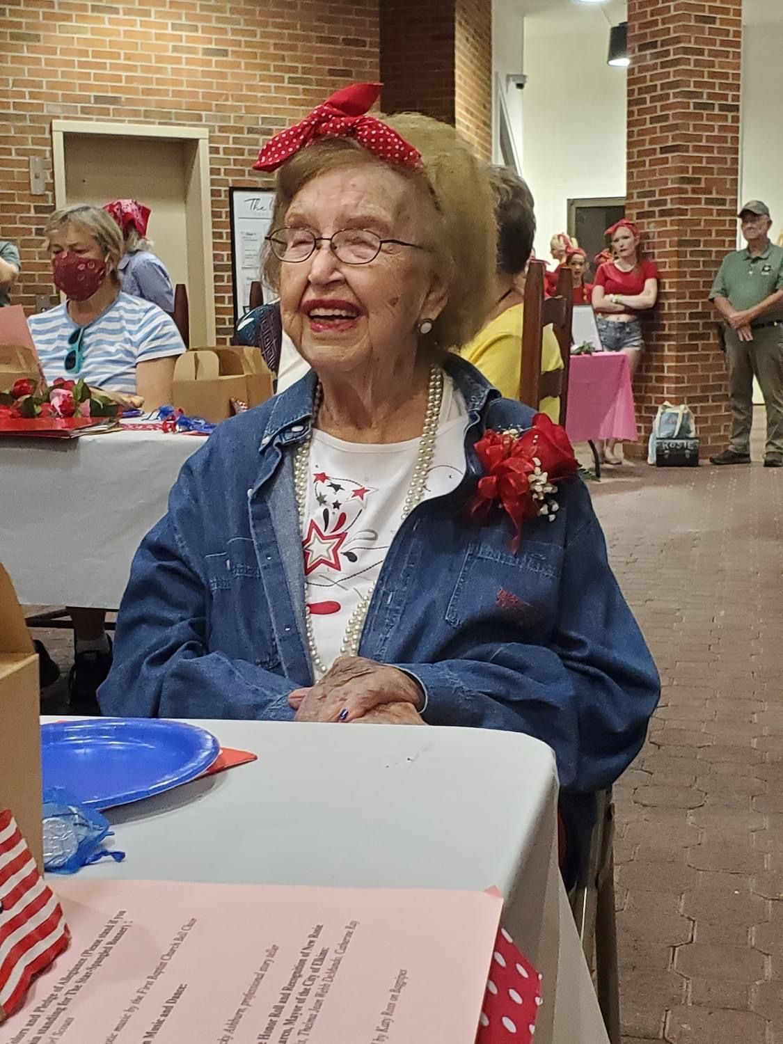 Elderly woman in denim jacket, red polka dot headband, smiling at an event.