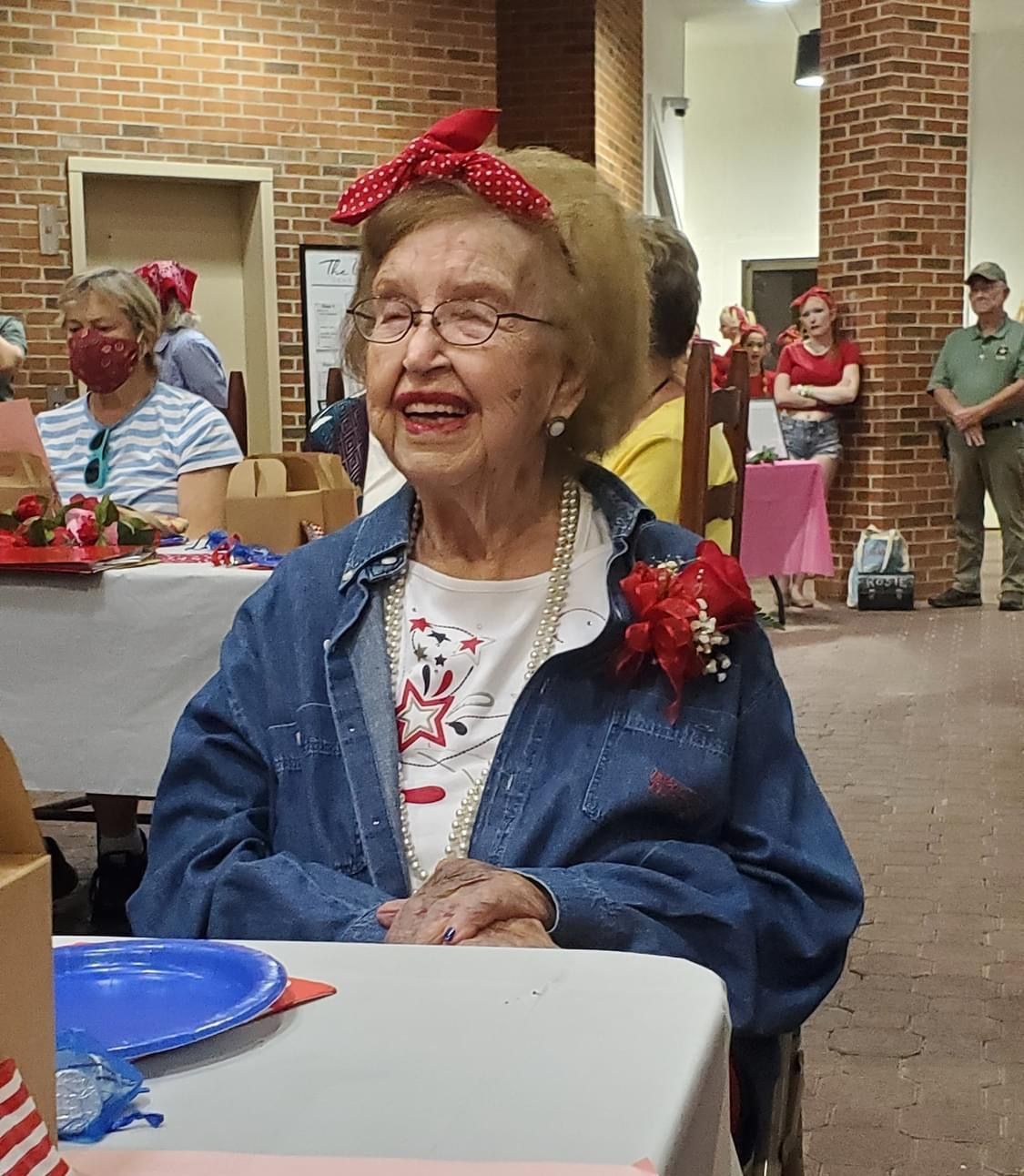 Smiling elderly woman wearing denim jacket and red bandana at an event; brick building background.