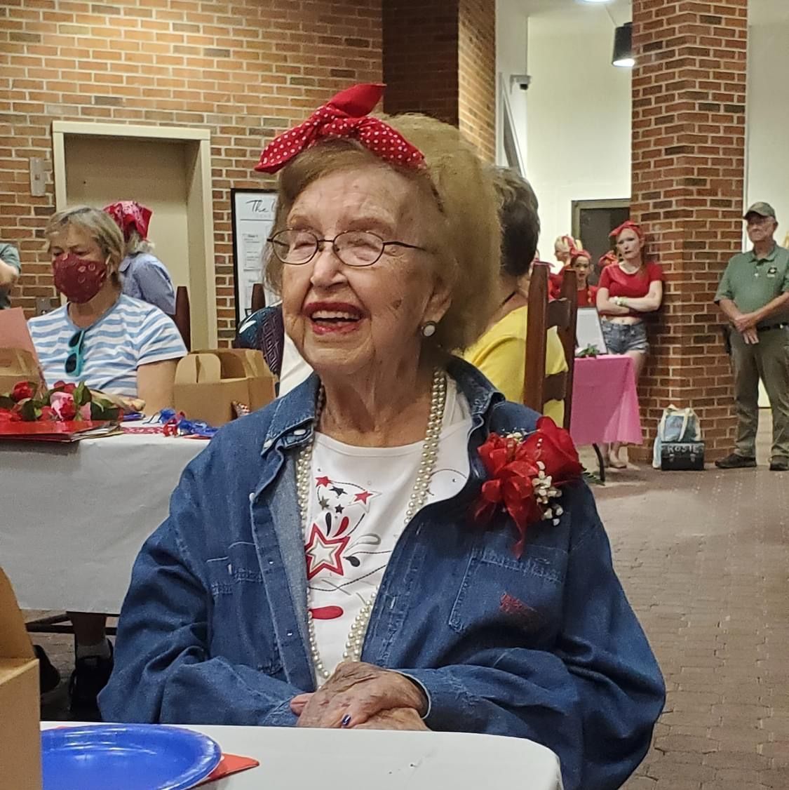 Elderly woman with a red bandana, denim jacket, and pearls, smiling at an event; other people in background.