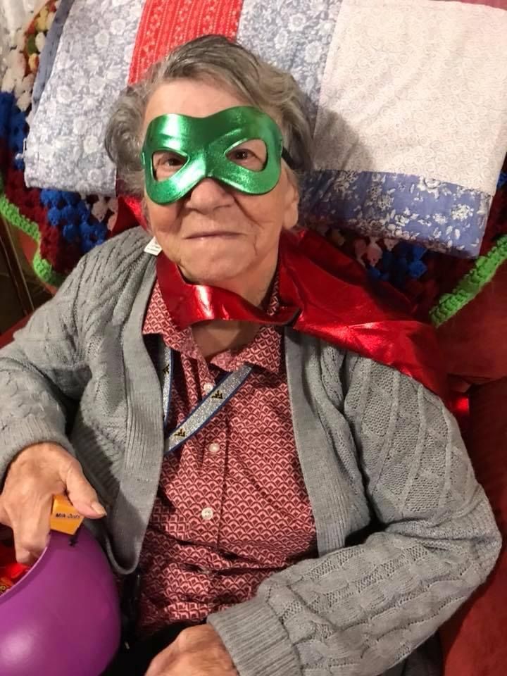Elderly woman in a green mask and red cape, holding a purple bucket, sitting.