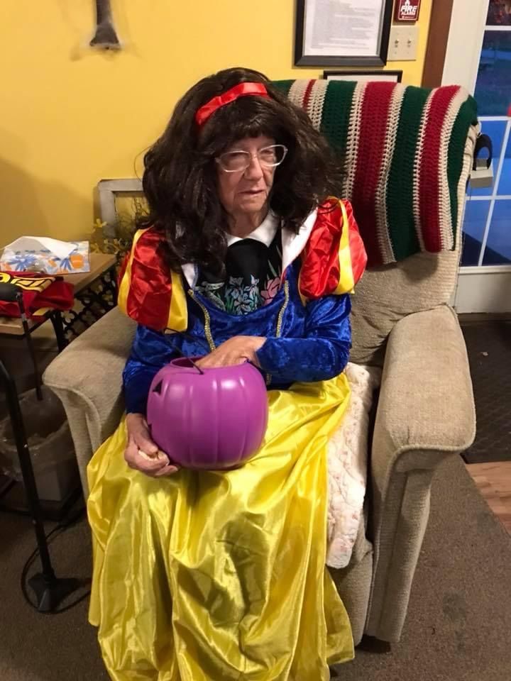 Person dressed as Snow White sitting in a chair, holding a purple pumpkin, wearing glasses.